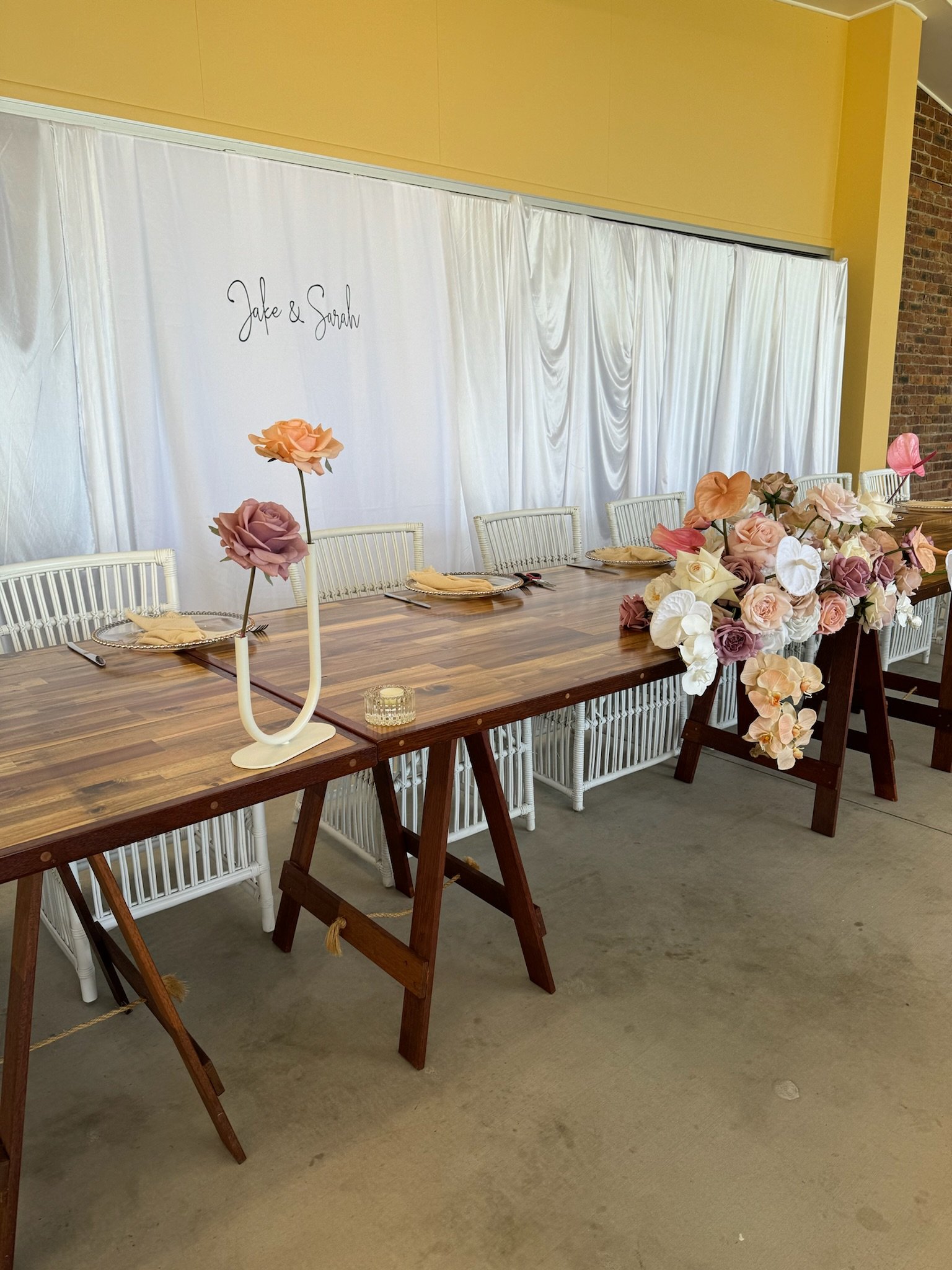 Decorative wedding table setup with a long wooden table, floral arrangements, white chairs, and a white backdrop with handwritten "Jake & Sarah" at a venue with yellow walls and brick accents.