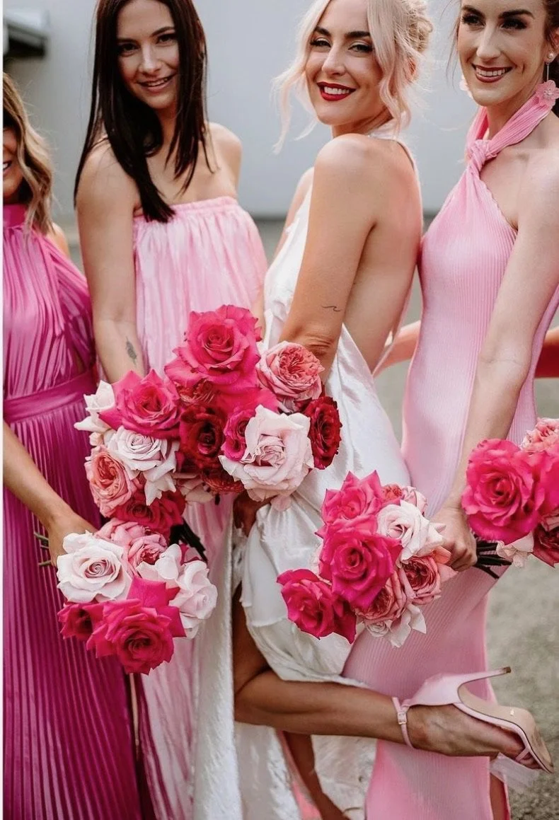 A group of women dressed in pink and white dresses, holding bouquets of pink and white roses, celebrating at a wedding or special event.