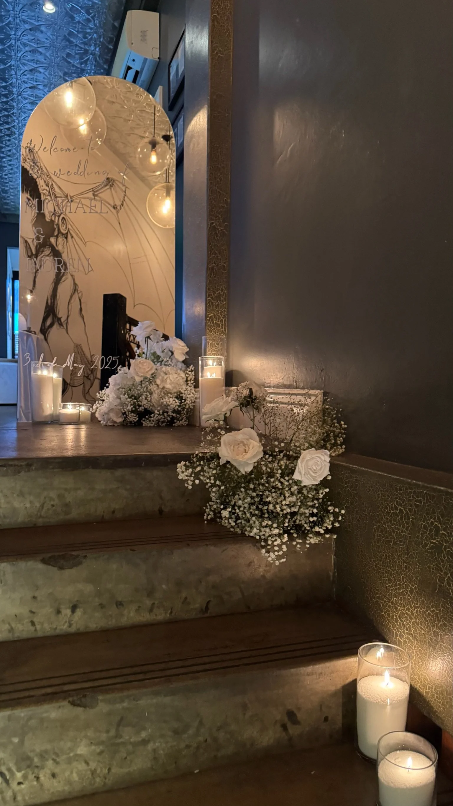 Wedding decoration with white flowers, candles, and a mirror with a drawing of a fairy and a welcome message for the wedding of Michael and Feren on May 3, 2025.