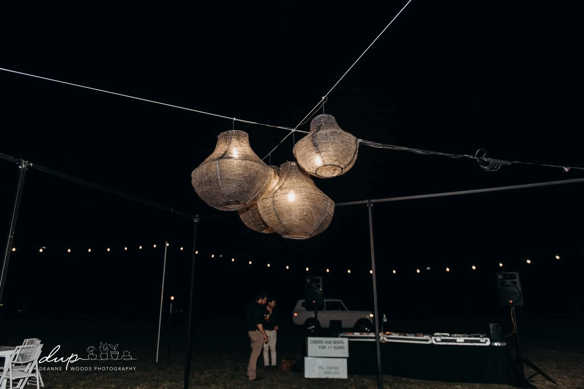 Night outdoor event with hanging woven pendant lights, two people talking near a sign, a DJ setup, and a vehicle in the background, under string lights.