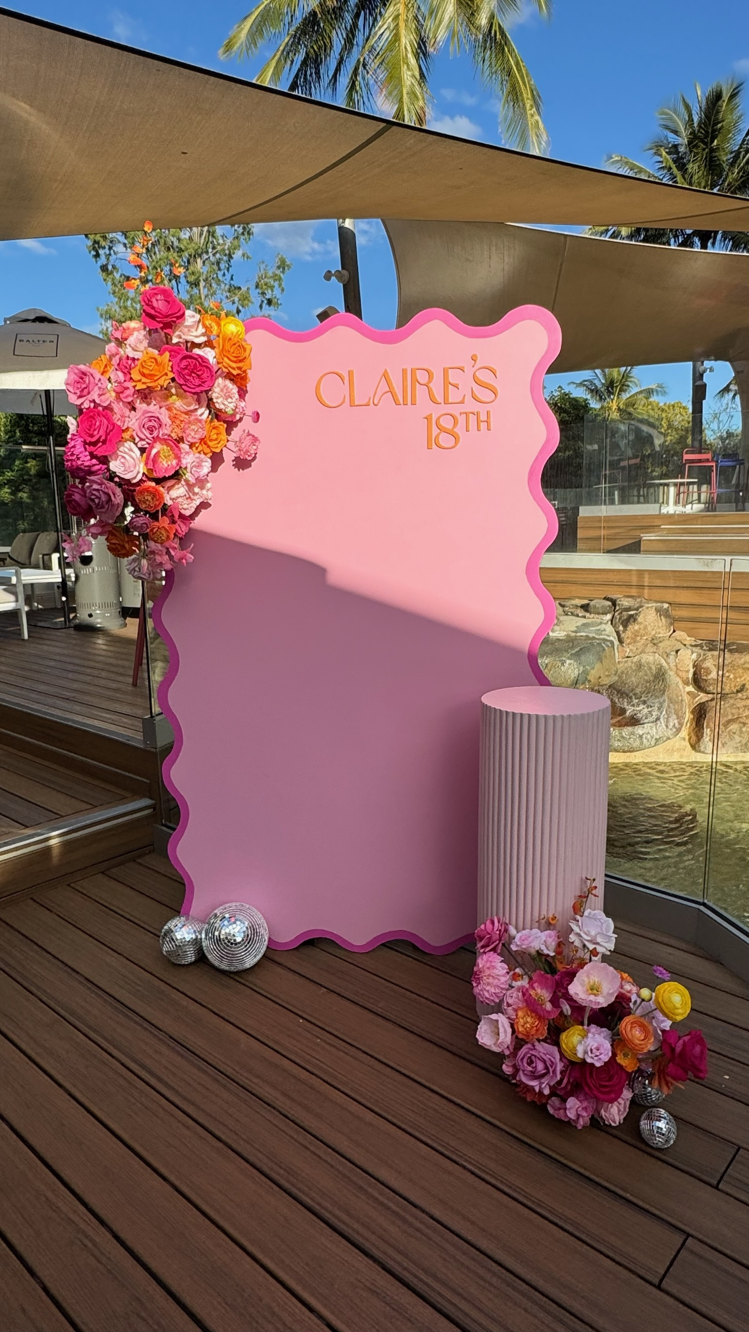 Pink birthday backdrop with flowers and disco balls, reading 'Claire's 18th' in gold lettering, set on a wooden deck under shade sails, with palm trees and water in the background.