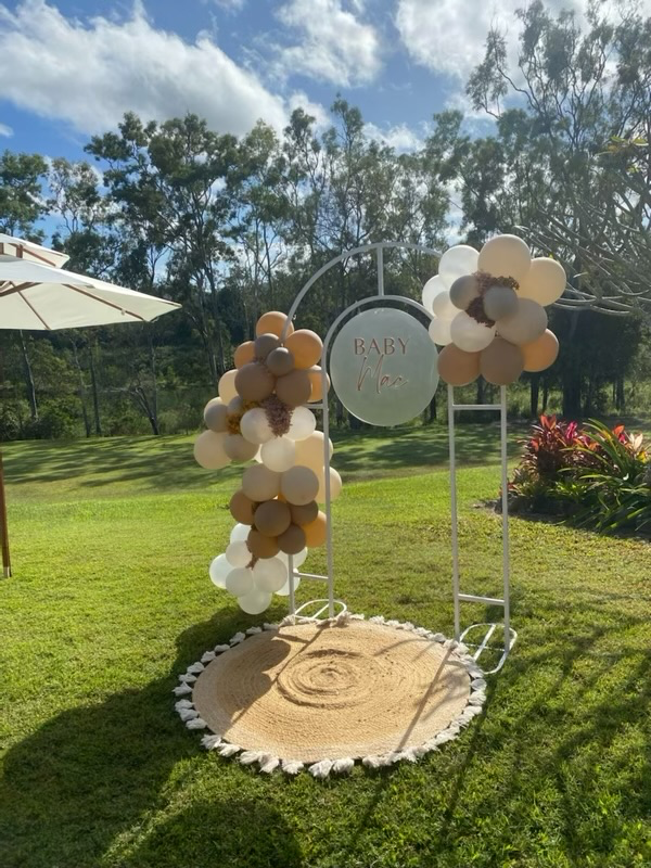 Outdoor celebration setup with a white metal arch decorated with balloons in neutral colors, on a grassy area with trees in the background. A round sign reads "Baby Moo" in the center of the arch. There is a circular rug with tassels on the grass und