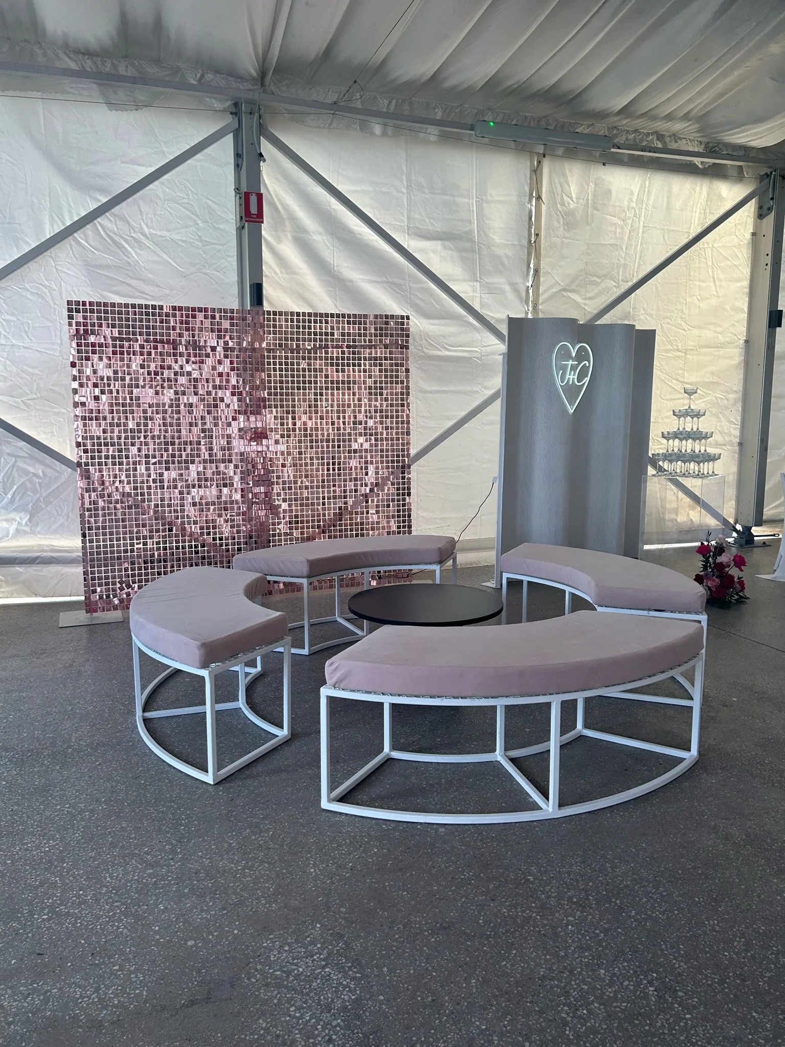 A wedding or event setup inside a white tent with modern furniture including curved benches with pastel pink cushions and a small round black table. In the background, there's a pixelated photo mosaic of a couple, a light-up heart with initials, a sm