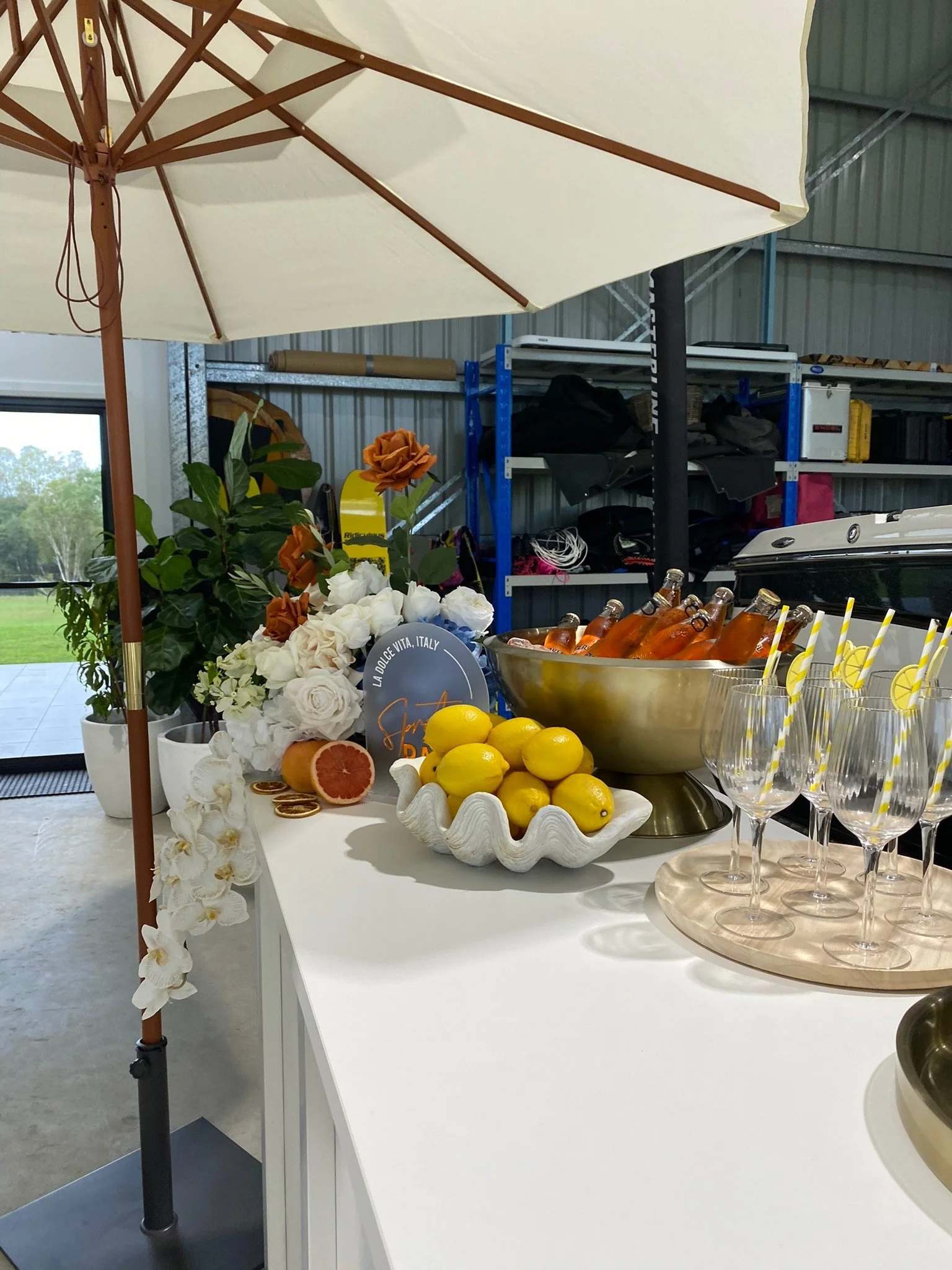 Bar table with lemon and grapefruit, champagne bottles in an ice bucket, empty champagne glasses with lemon slices, and floral decorations under a large patio umbrella.