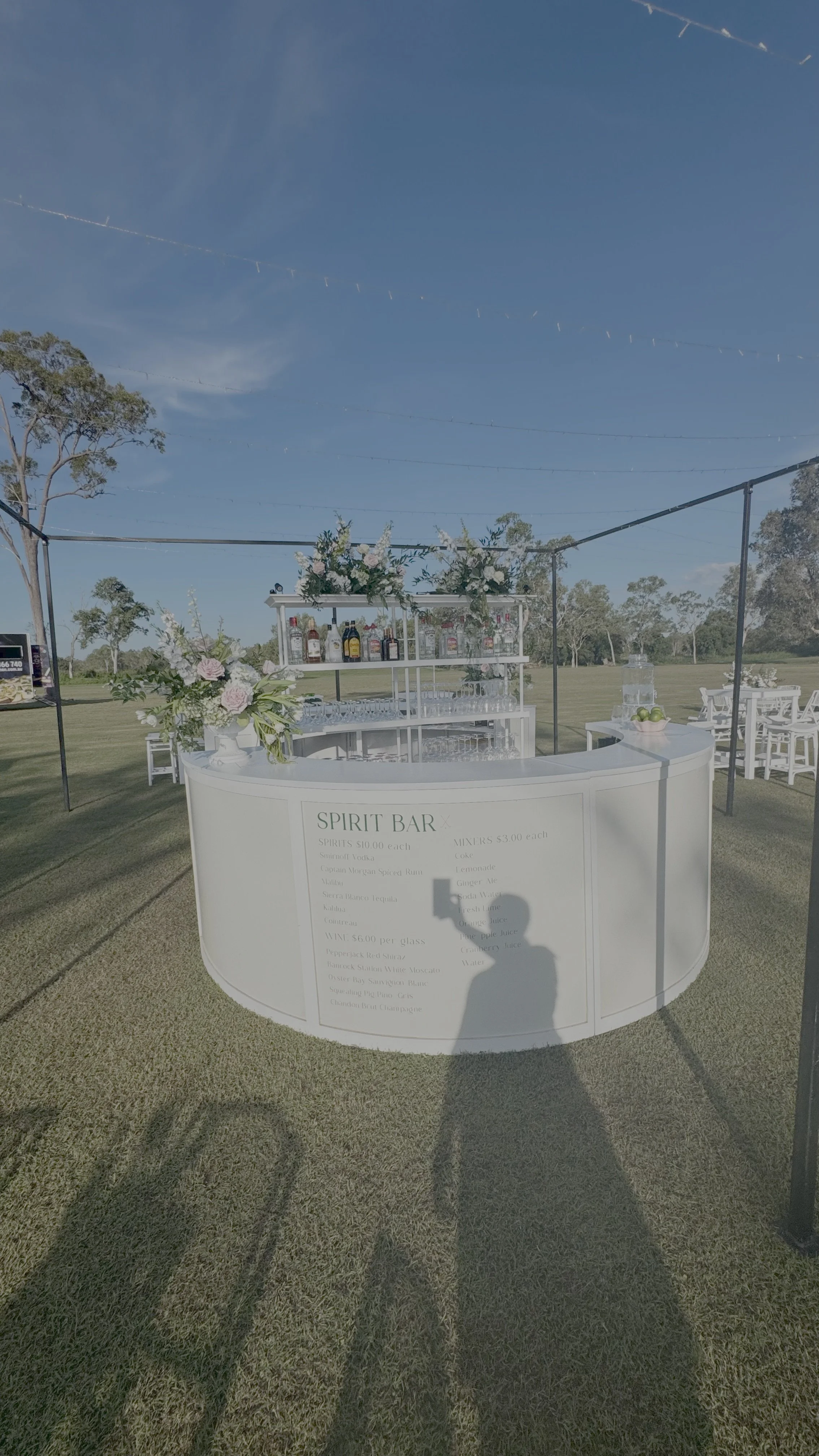 Outdoor bar setup with floral arrangements and liquor bottles on a sunny day, surrounded by grass and trees.
