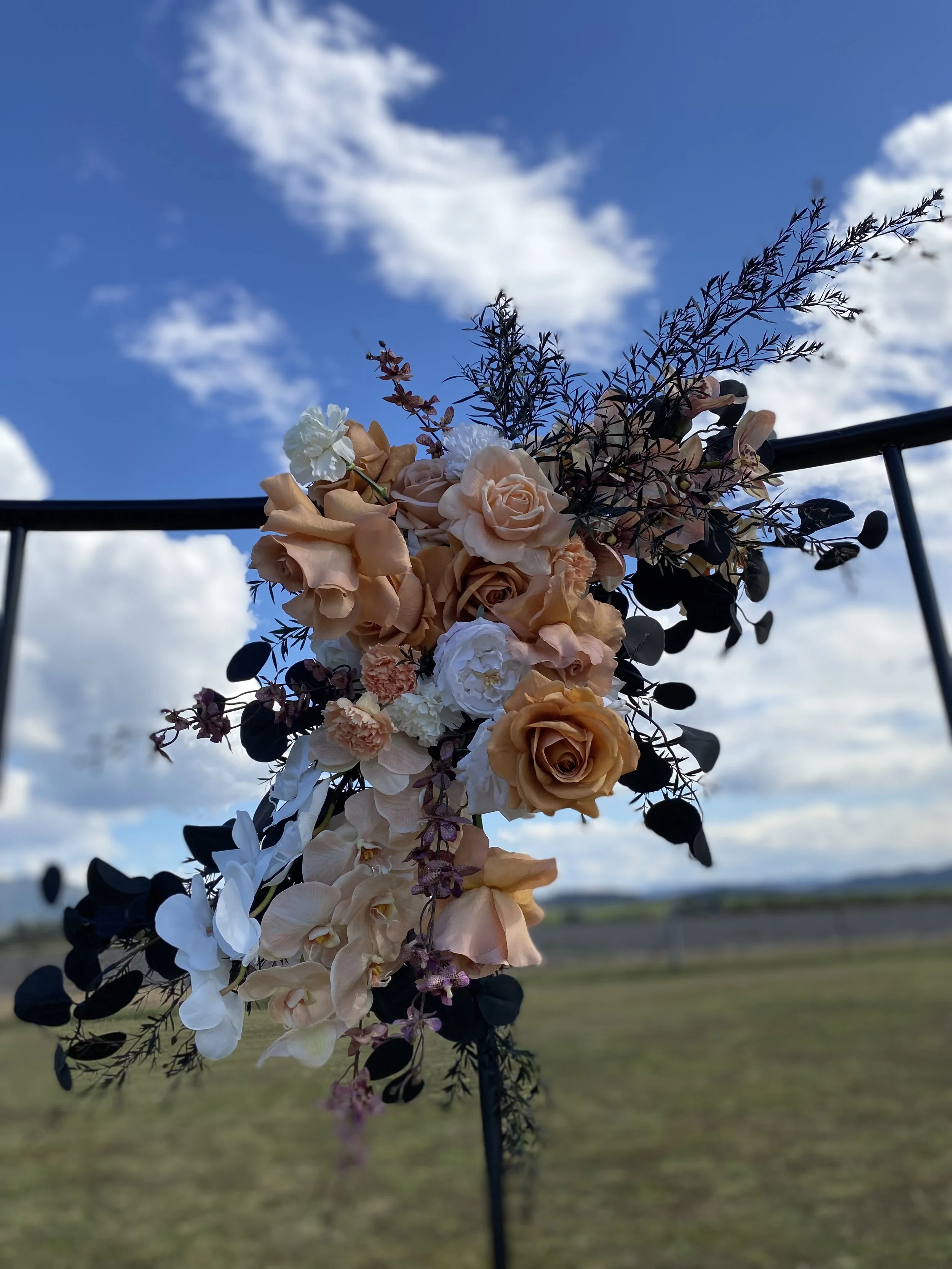 A floral arrangement with peach, white, and purple flowers hanging from a black metal arch against a blue sky with clouds.