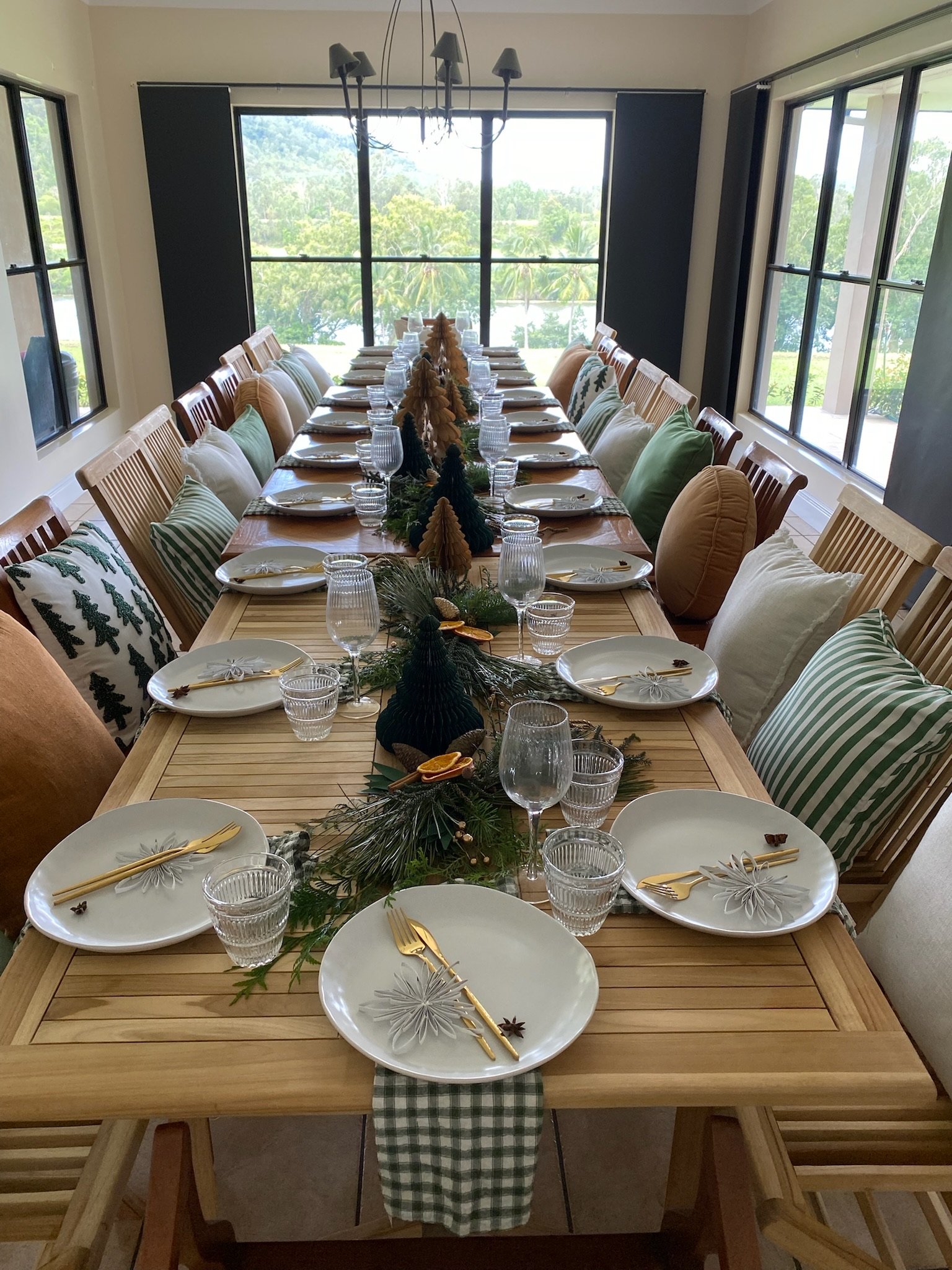 A long wooden dining table set for a holiday meal with plates, gold utensils, wine glasses, and green and white striped and Christmas-themed pillows on wooden chairs. The table has a Christmas centerpiece with greenery, pinecones, and small decorativ