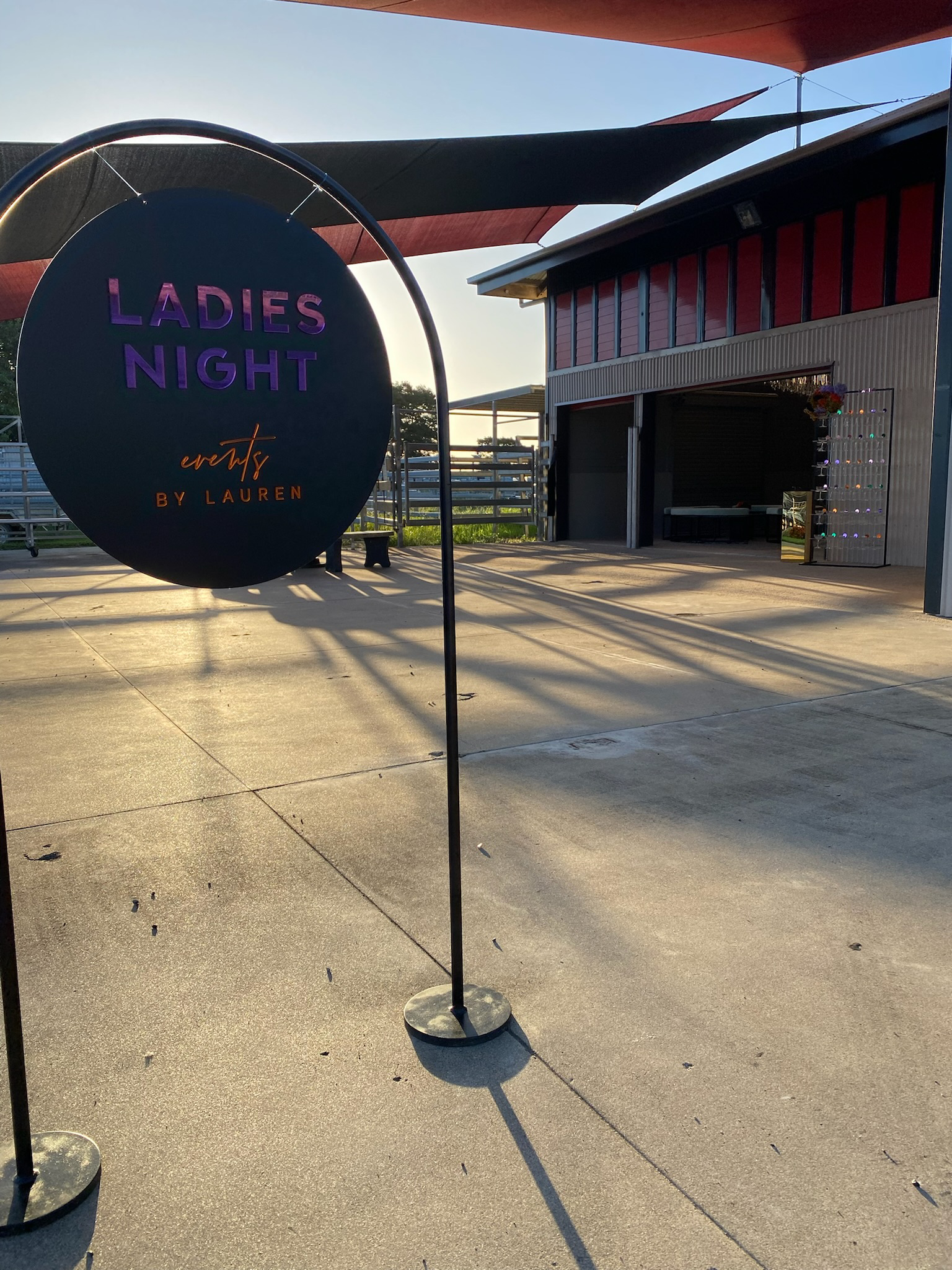 A sign reading 'Ladies Night' at an outdoor event, with a barn-like building in the background and shade sails overhead.