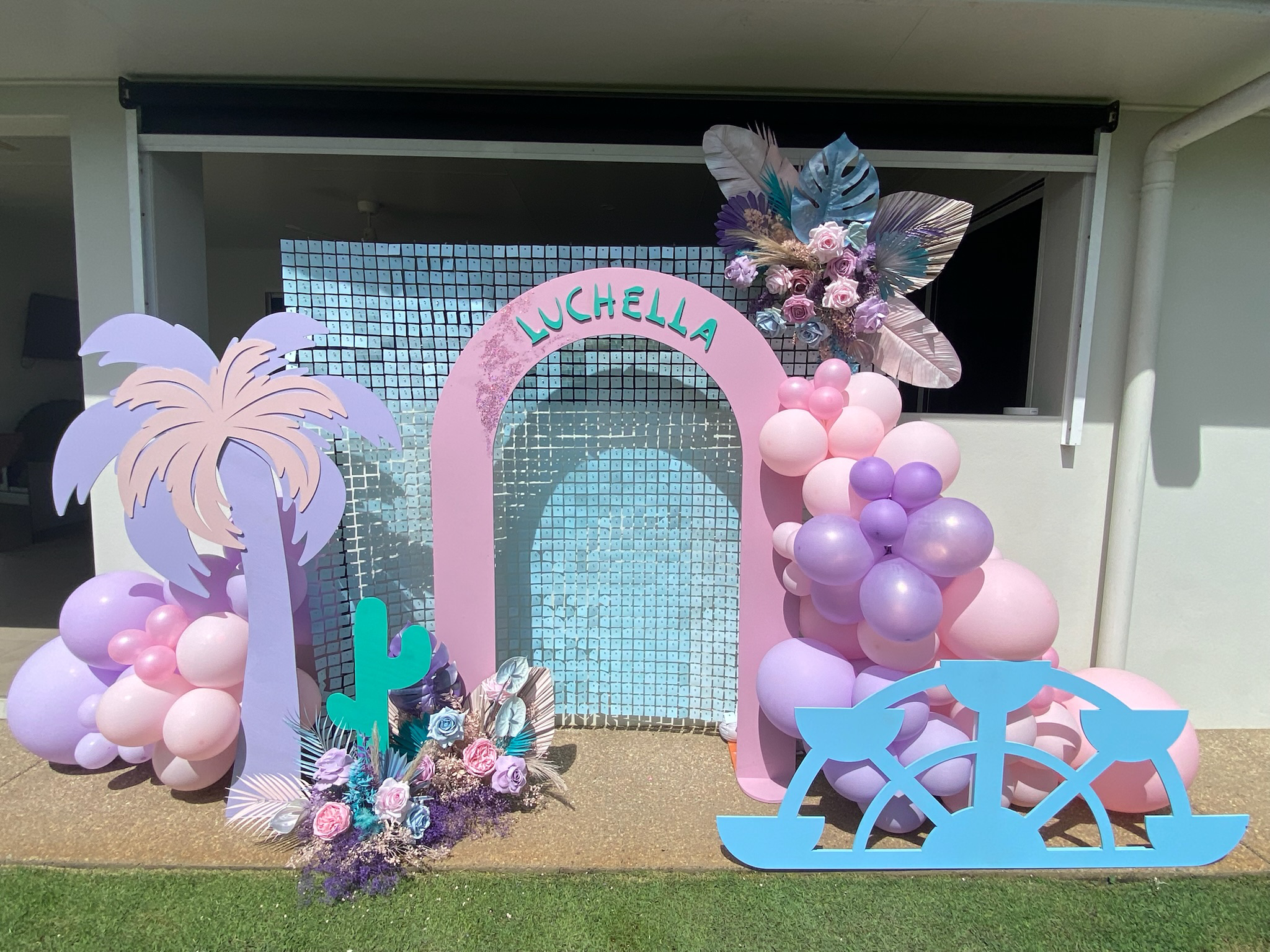 Decorative display with pink and purple balloons, a pink arch with the name 'Luchella', floral arrangements, paper palm trees, a cactus cutout, and a blue sailboat cutout, set up outdoors.