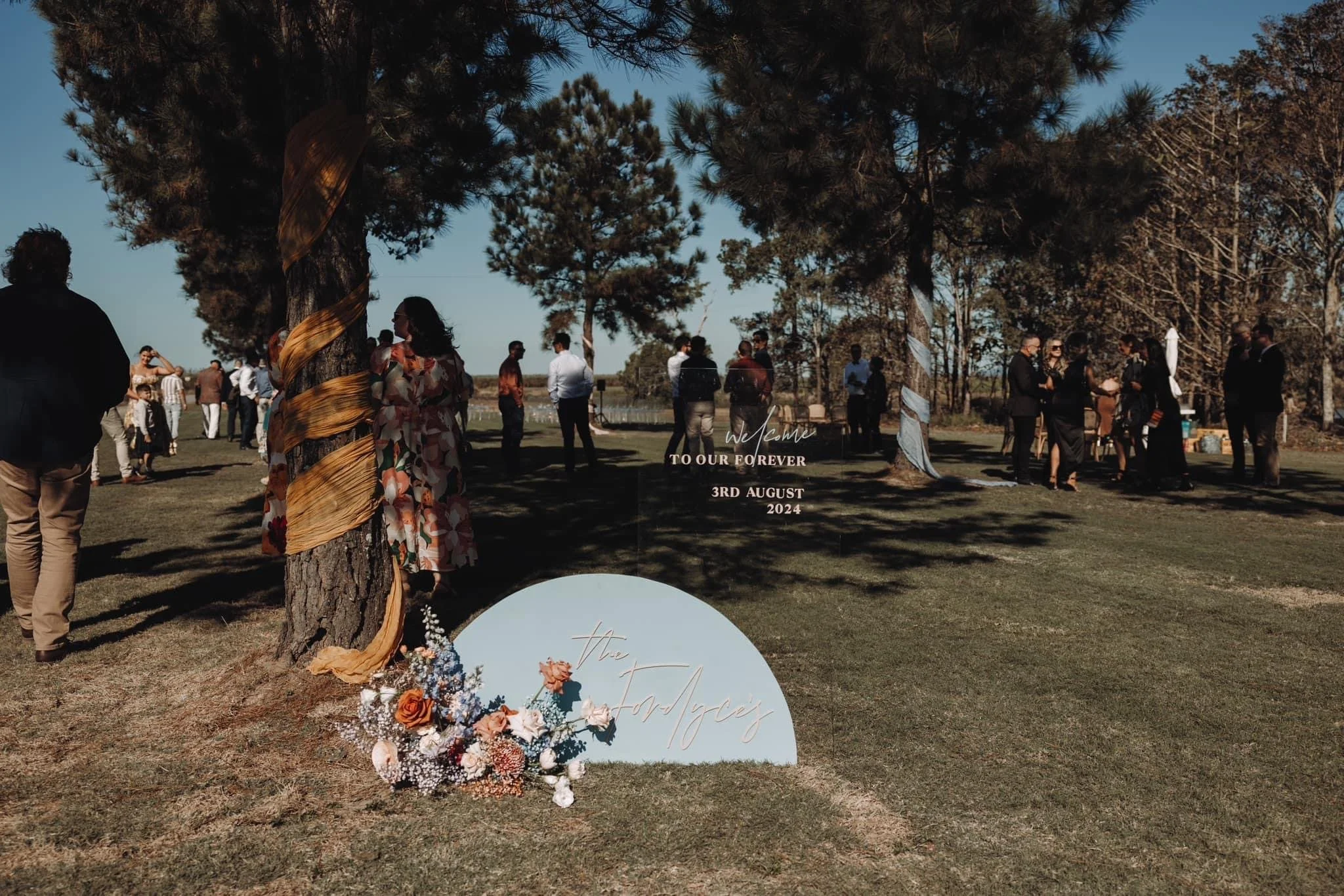Outdoor wedding celebration with guests mingling on a grassy area under large trees, with decorative signs and floral arrangements, on a sunny day.