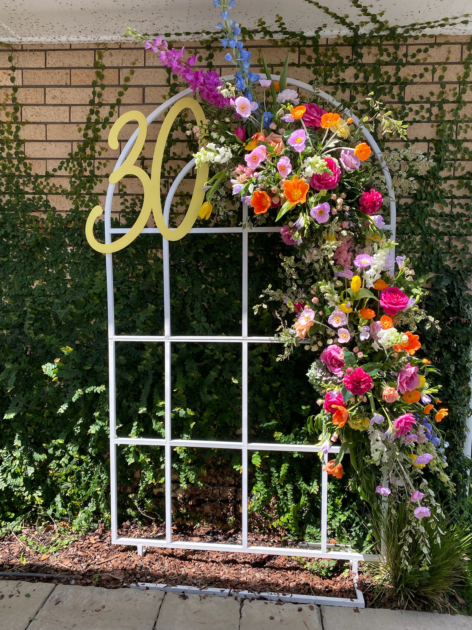 A decorative white metal arbor decorated with colorful flowers and the number 30 in yellow, set against a brick wall with vines.