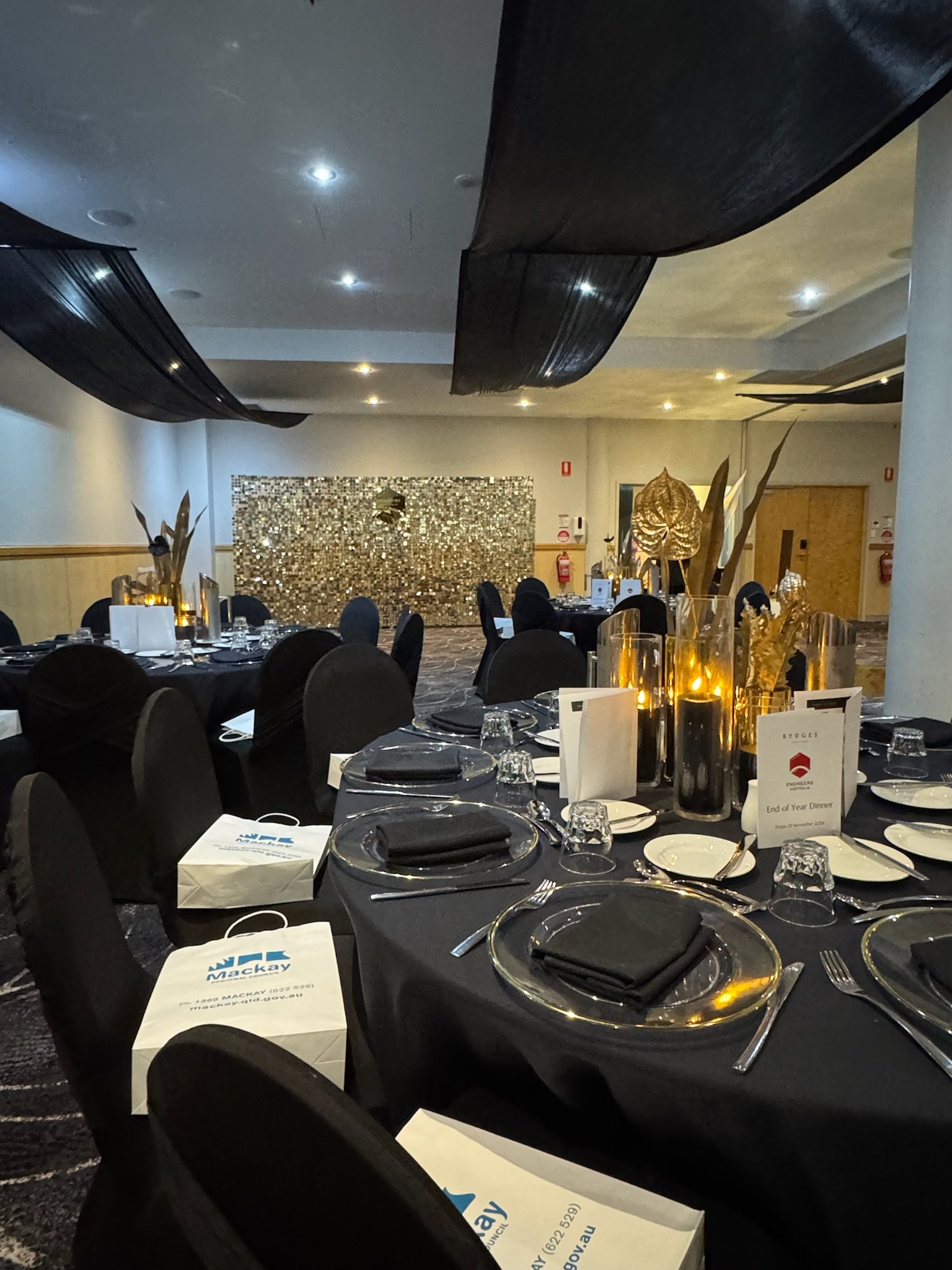 Decorated banquet hall with round tables set with black napkins, glassware, and candles, gold and black accents, and a textured wall in the background.