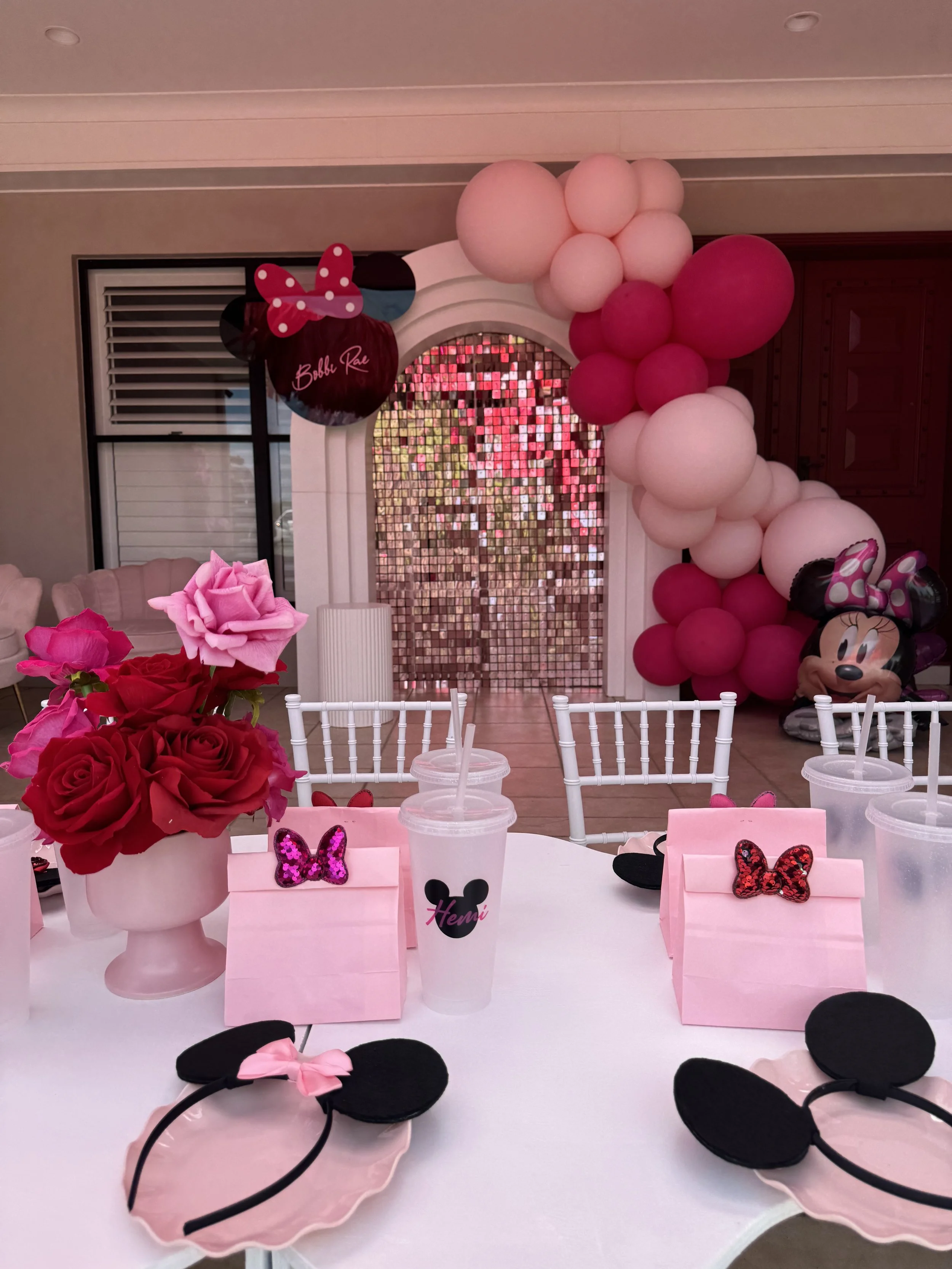 Party table decorated with pink and black Minnie Mouse theme, featuring Minnie Mouse headbands, pink and red roses in a vase, pink gift bags with Minnie Mouse bows, and a large balloon arch with pink and white balloons behind a pink sequin backdrop.