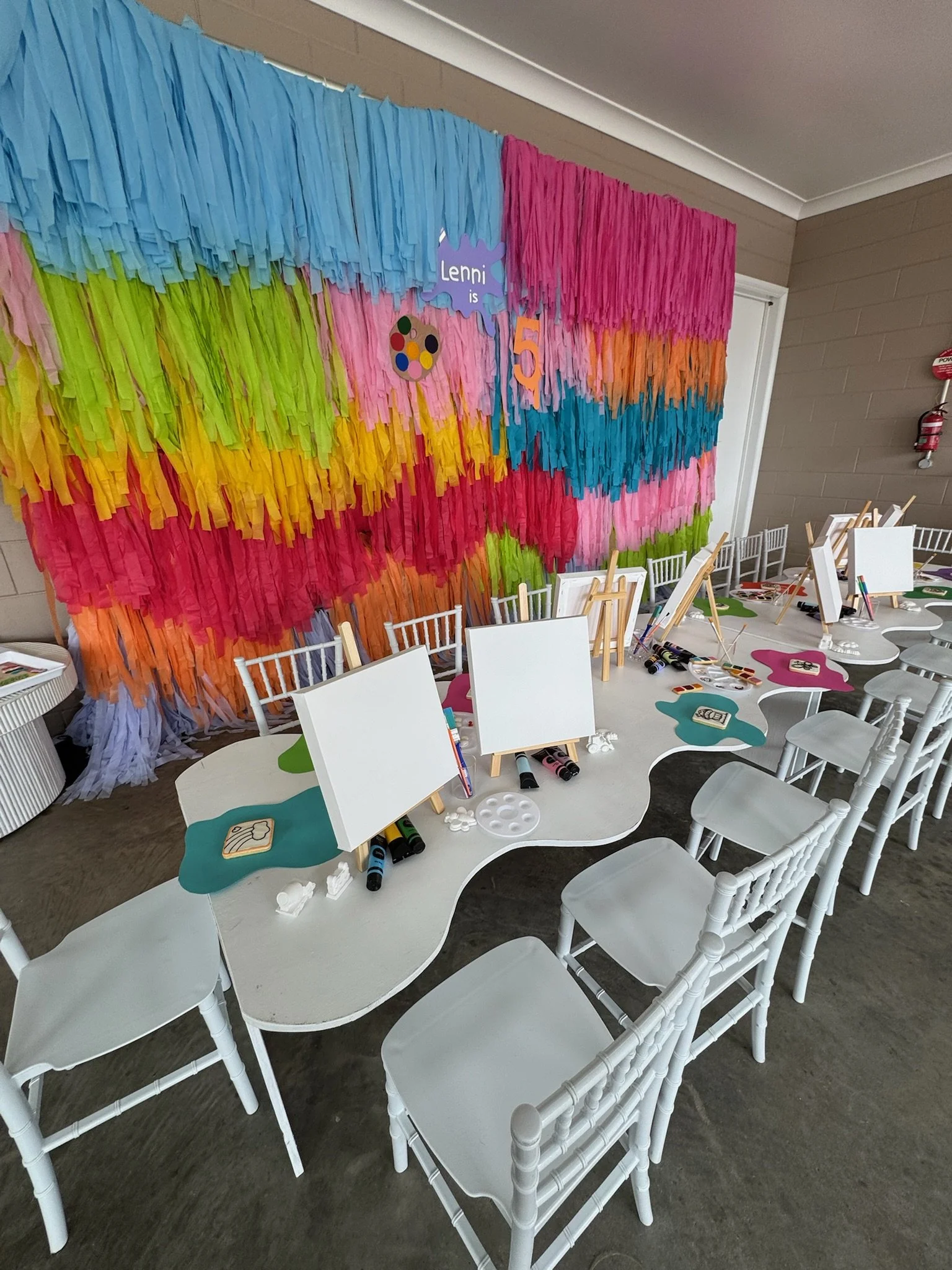 Colorful birthday party setup with a rainbow paper backdrop, small easels, painting supplies, and a table with seating for kids.