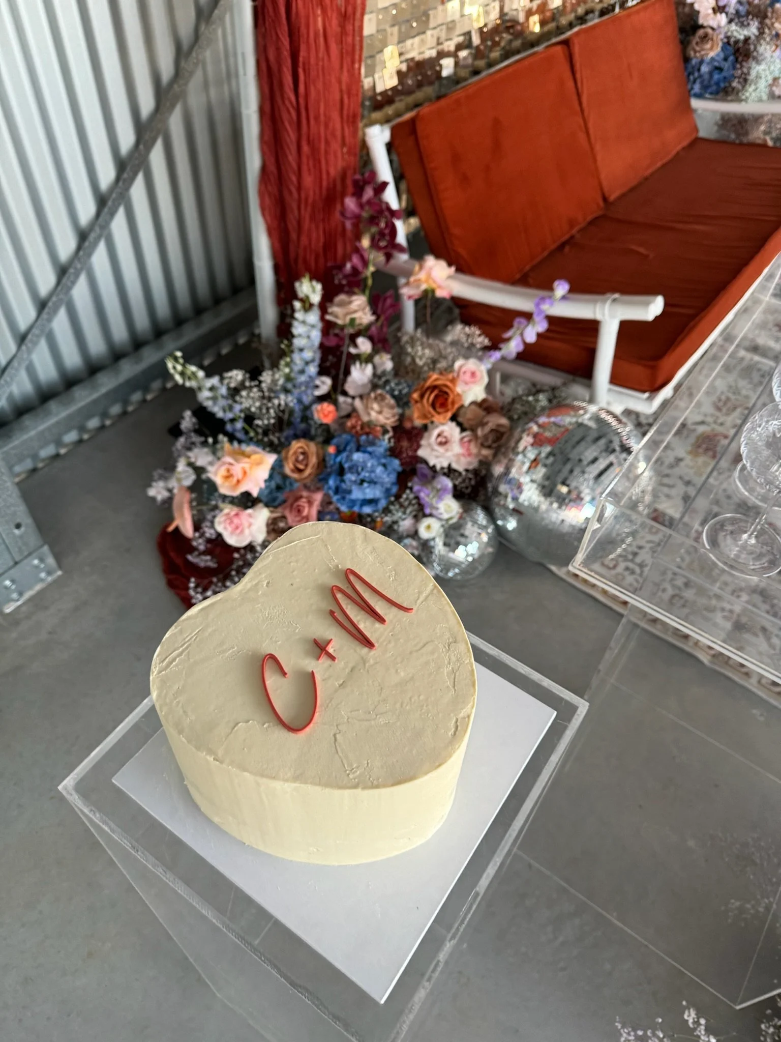 Heart-shaped cake with red writing on top, placed on a transparent stand. Behind it, a bouquet of colorful flowers, a red sofa, mirrored disco balls, and decorative items are visible.