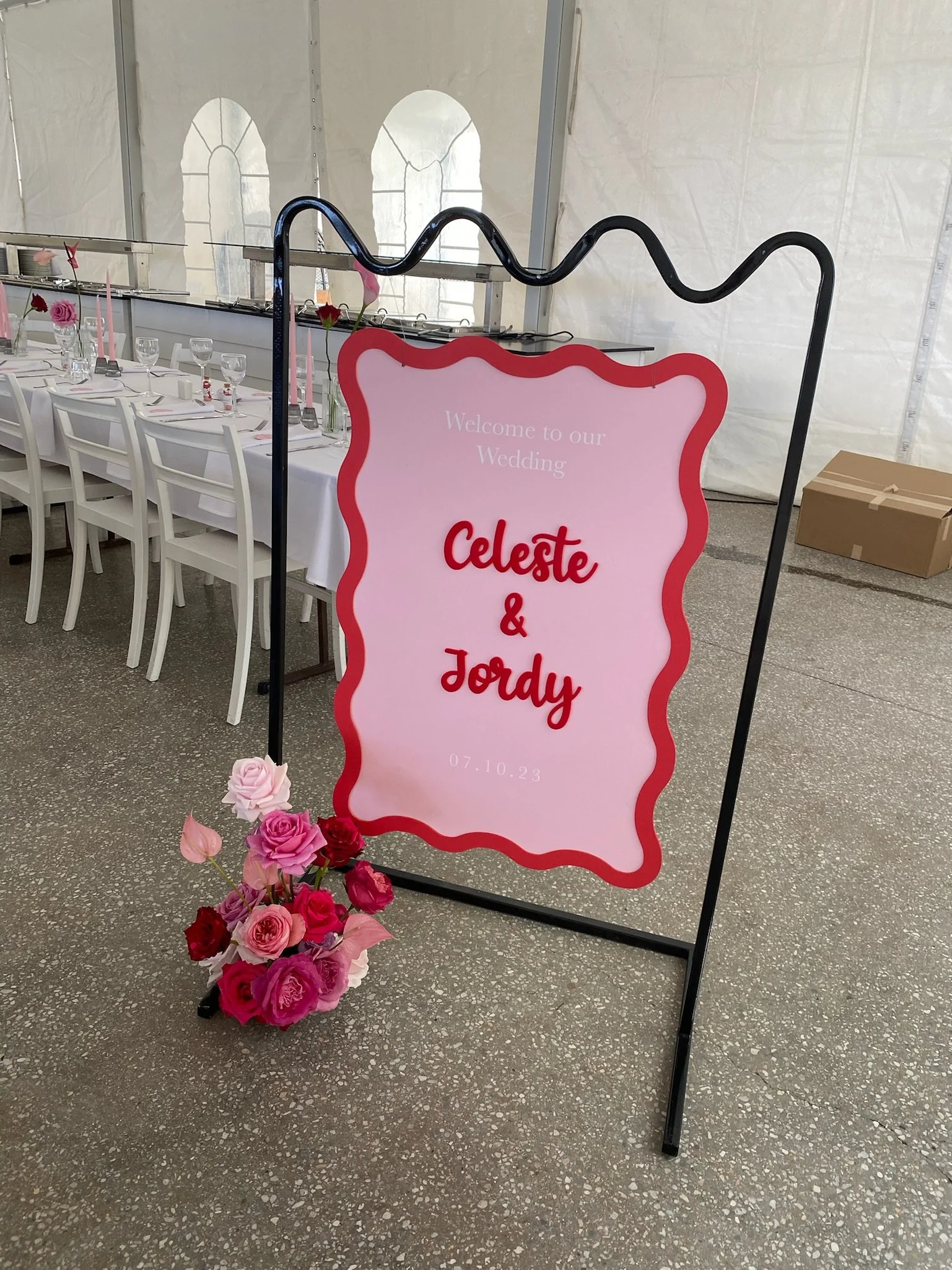 Wedding welcome sign with pink background and red border, displaying the names Celeste and Jordy, date 07.10.23, surrounded by pink and red roses, in a white tent with decorated dining tables.