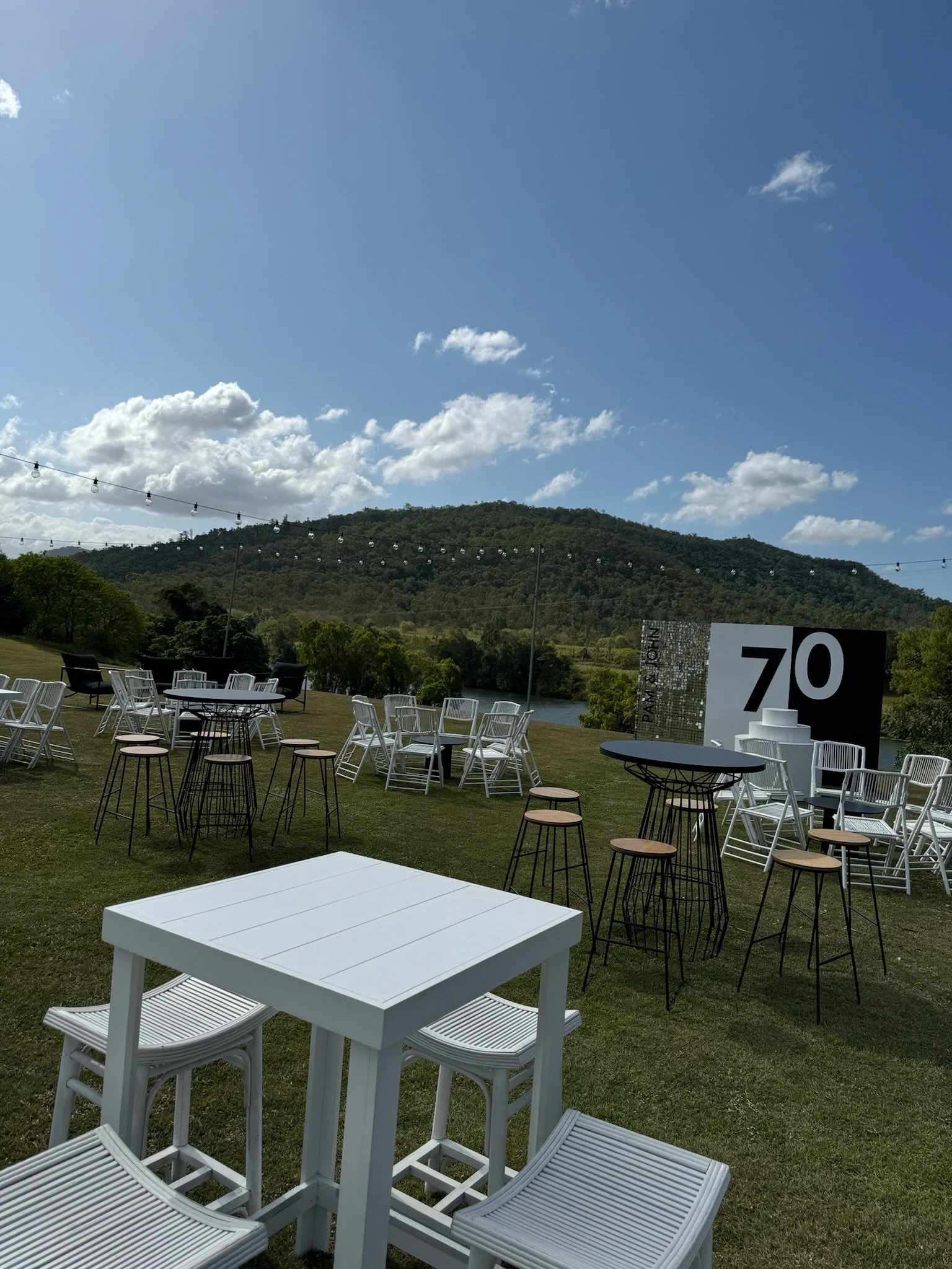 Outdoor event setup on a grassy area with tables, chairs, and string lights, near a body of water with hills in the background, under a partly cloudy sky, celebrating a 70th milestone.