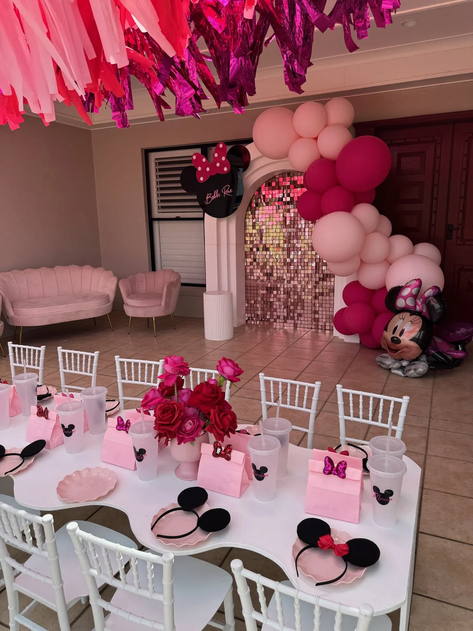 Party room decorated with pink and white balloons, a Minnie Mouse theme, pink roses in a vase, and Minnie Mouse ears on the table, indicating a Minnie Mouse-themed birthday celebration.