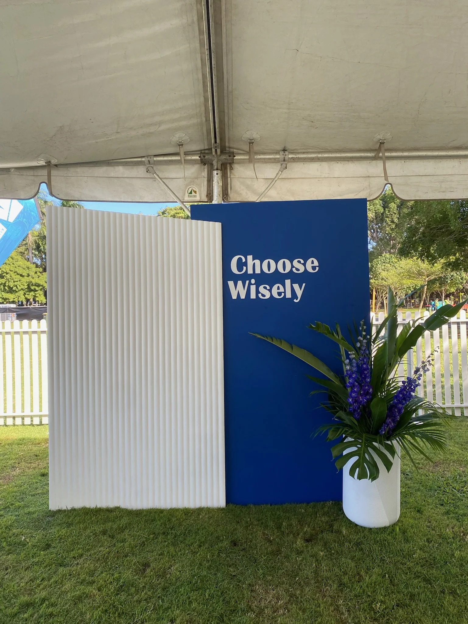 A blue sign with white text that says "Choose Wisely" is positioned next to a white corrugated panel and a white vase filled with purple flowers and green leafy plants, under a white tent outdoors.