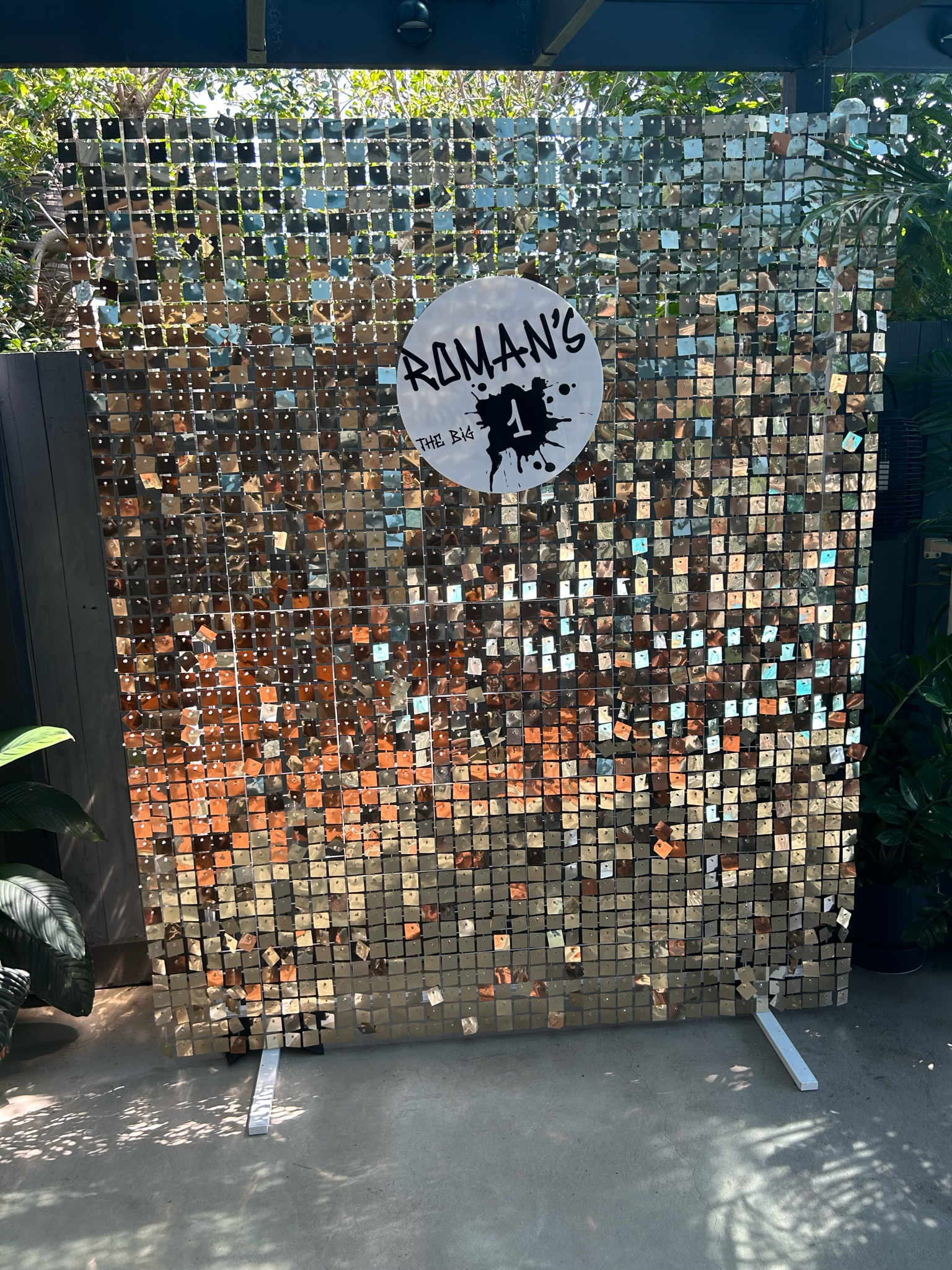 Decorative backdrop made of small, reflective tiles with a sign reading "ROMAN'S THE BIG 1" in the center.