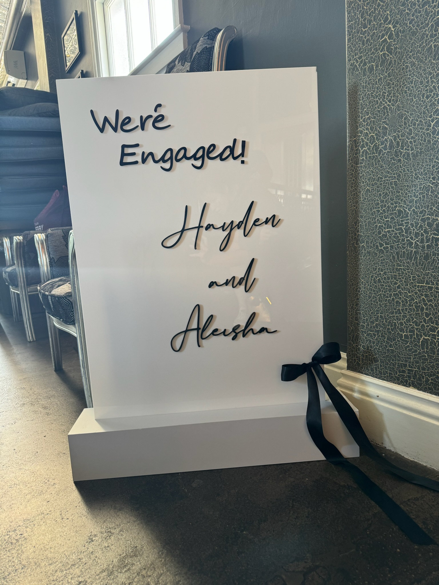 White sign with black text reading 'We're Engaged! Hayden and Aleasha' and a black ribbon tied to the bottom right corner, placed on the floor next to a wall with textured black and gold wallpaper, in a room with chairs and windows.