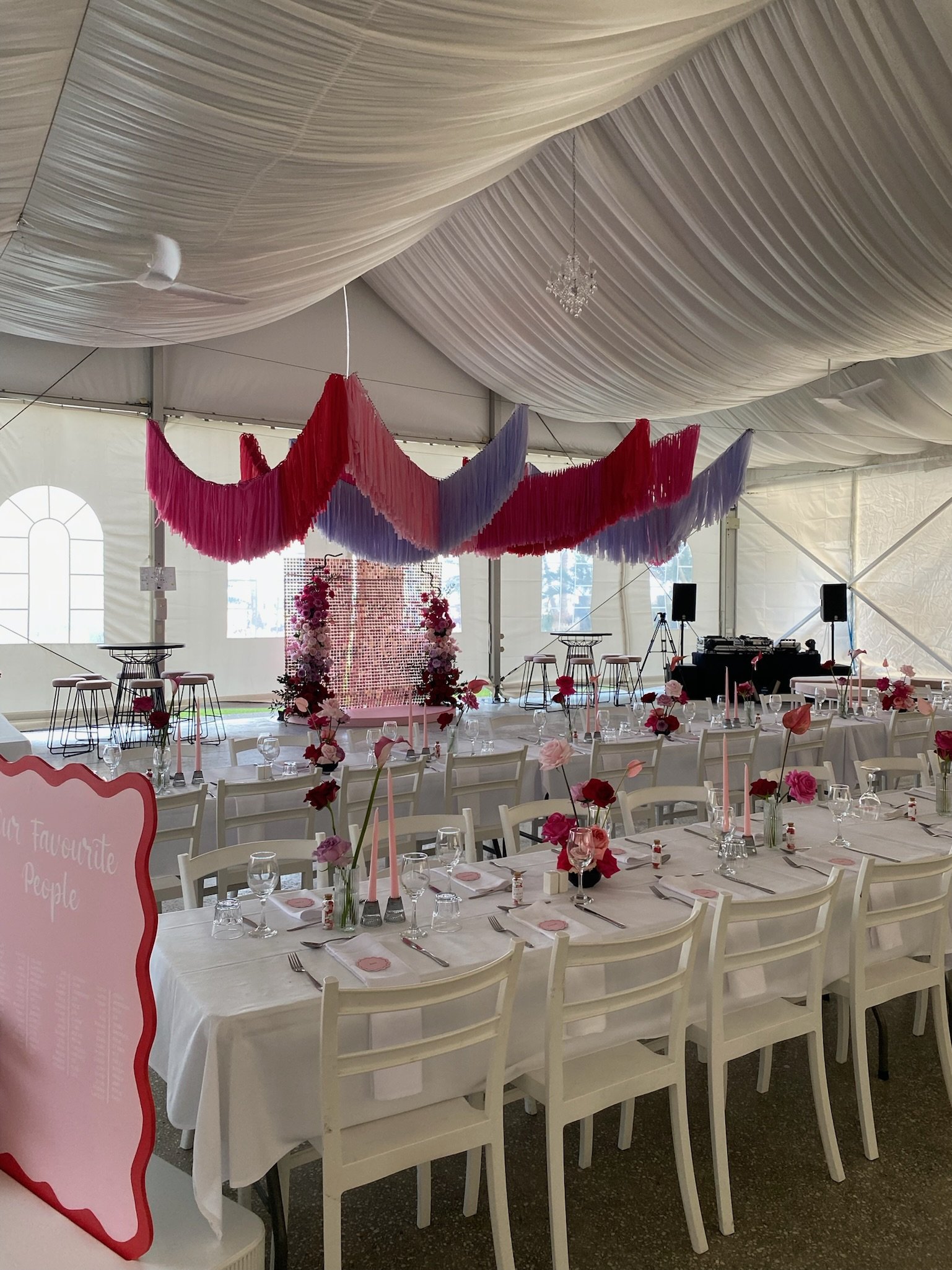 Decorated event tent with white tablecloths, pink and red flowers, pink candles, and pink napkins, pink and purple hanging fabric streamers, and a floral backdrop with pink and red flowers.
