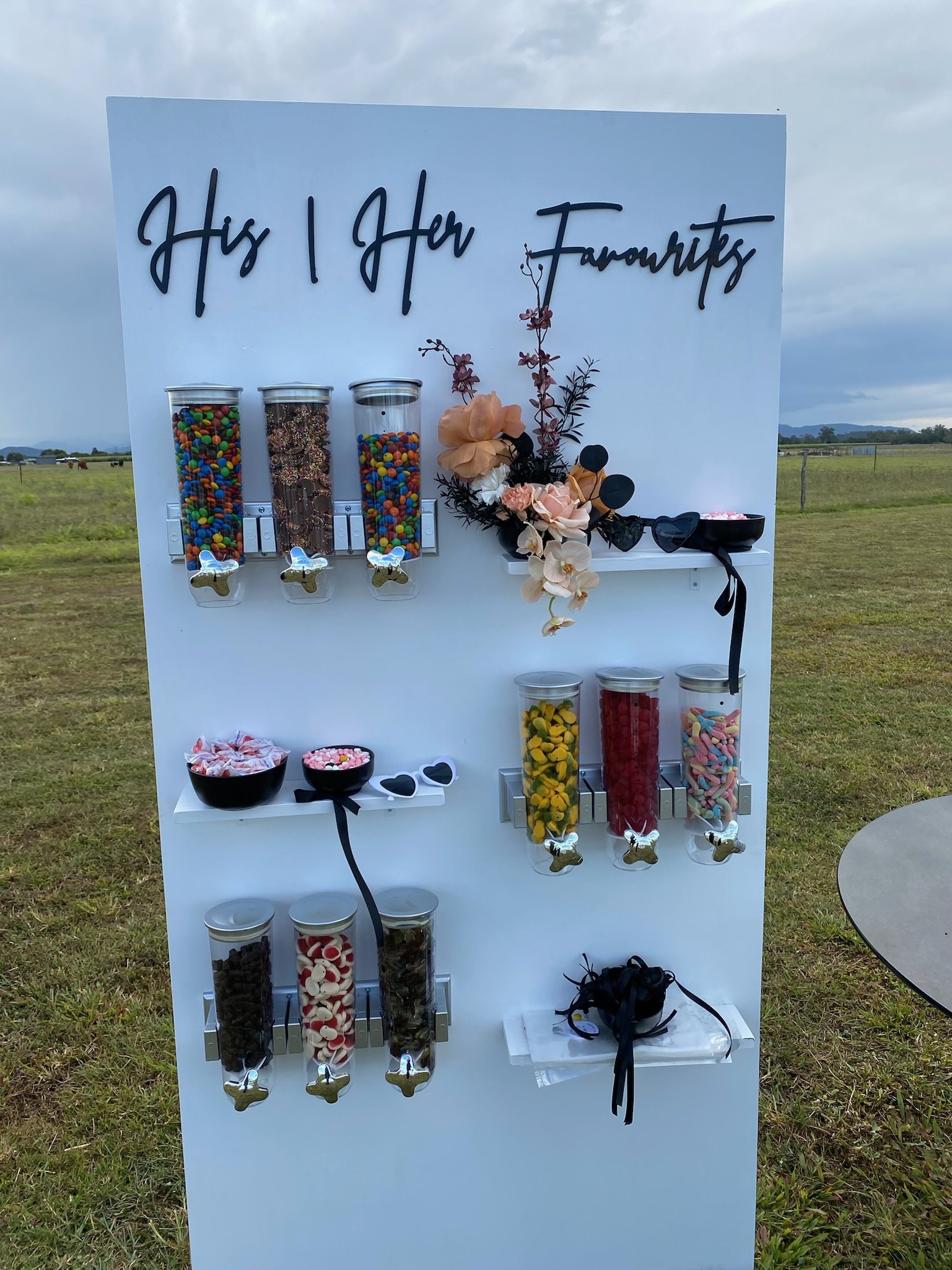 Candy buffet with jars of colorful candies, black bowls of wrapped sweets, and a sign that reads 'His & Her Favourites' with decorative flowers and sunglasses.