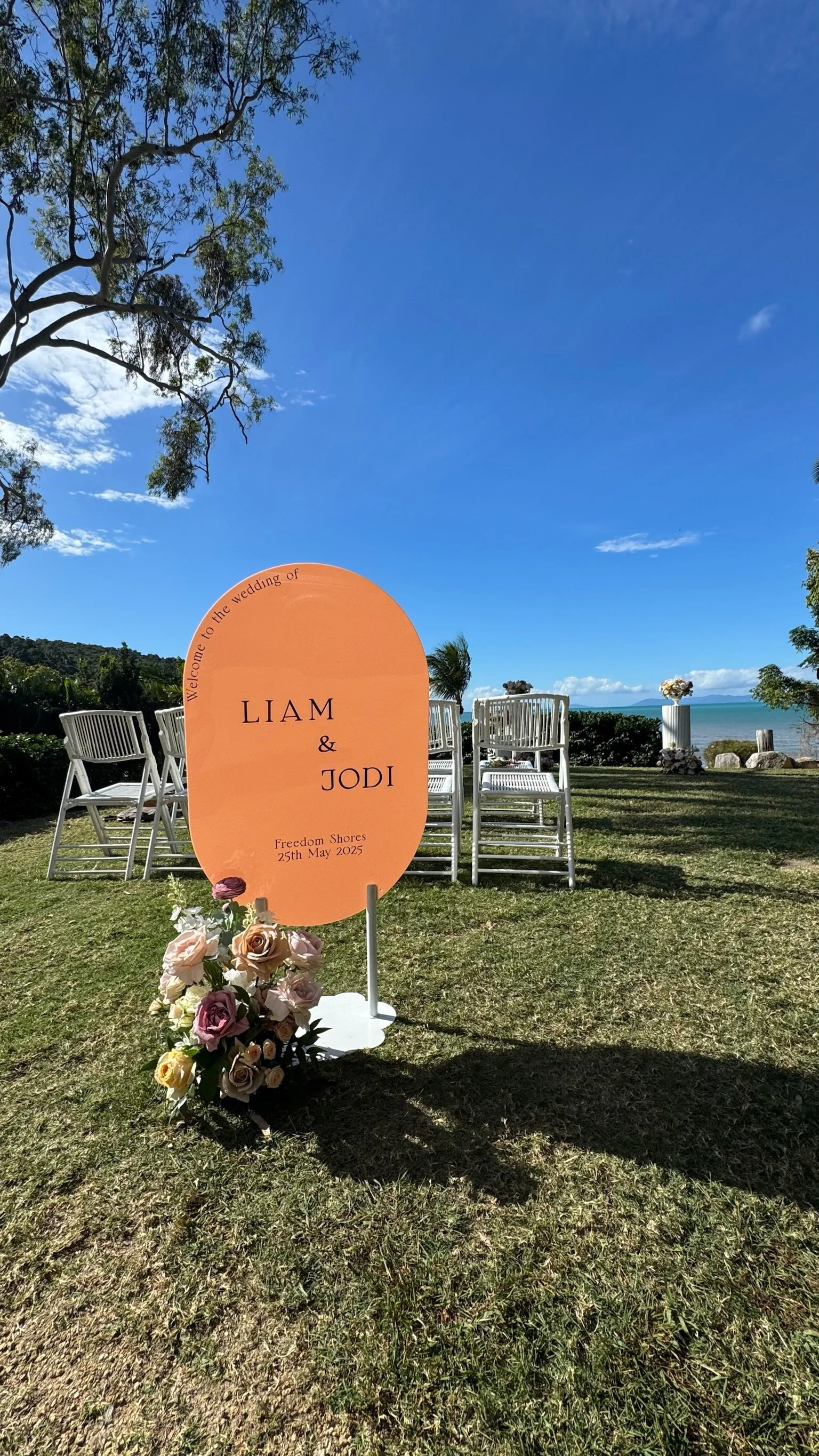 Wedding sign for Liam and Jodi at Freedom Shores on May 25, 2025, decorated with flowers, set outdoors on a grassy area with chairs and a view of the ocean in the background.