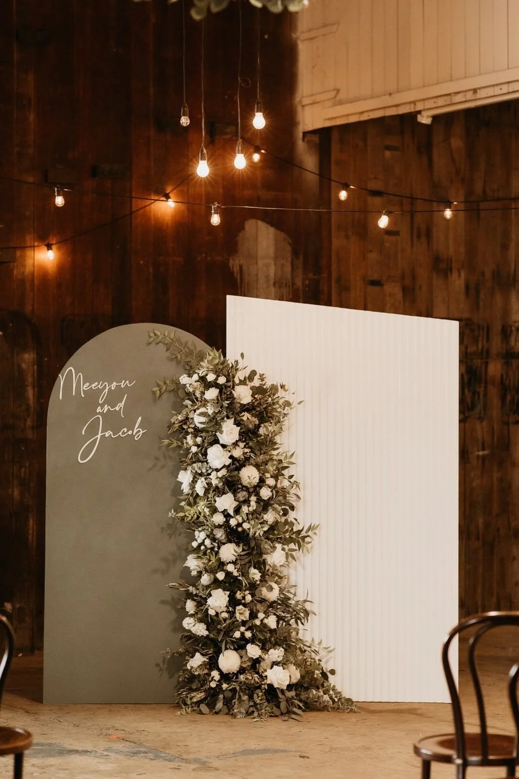 A wedding backdrop with a curved gray panel on the left edged with greenery and white flowers, and a plain white panel on the right. The gray panel has the handwritten names 'Meyson and Jacob.' The setting has warm ambient lighting from hanging bulbs