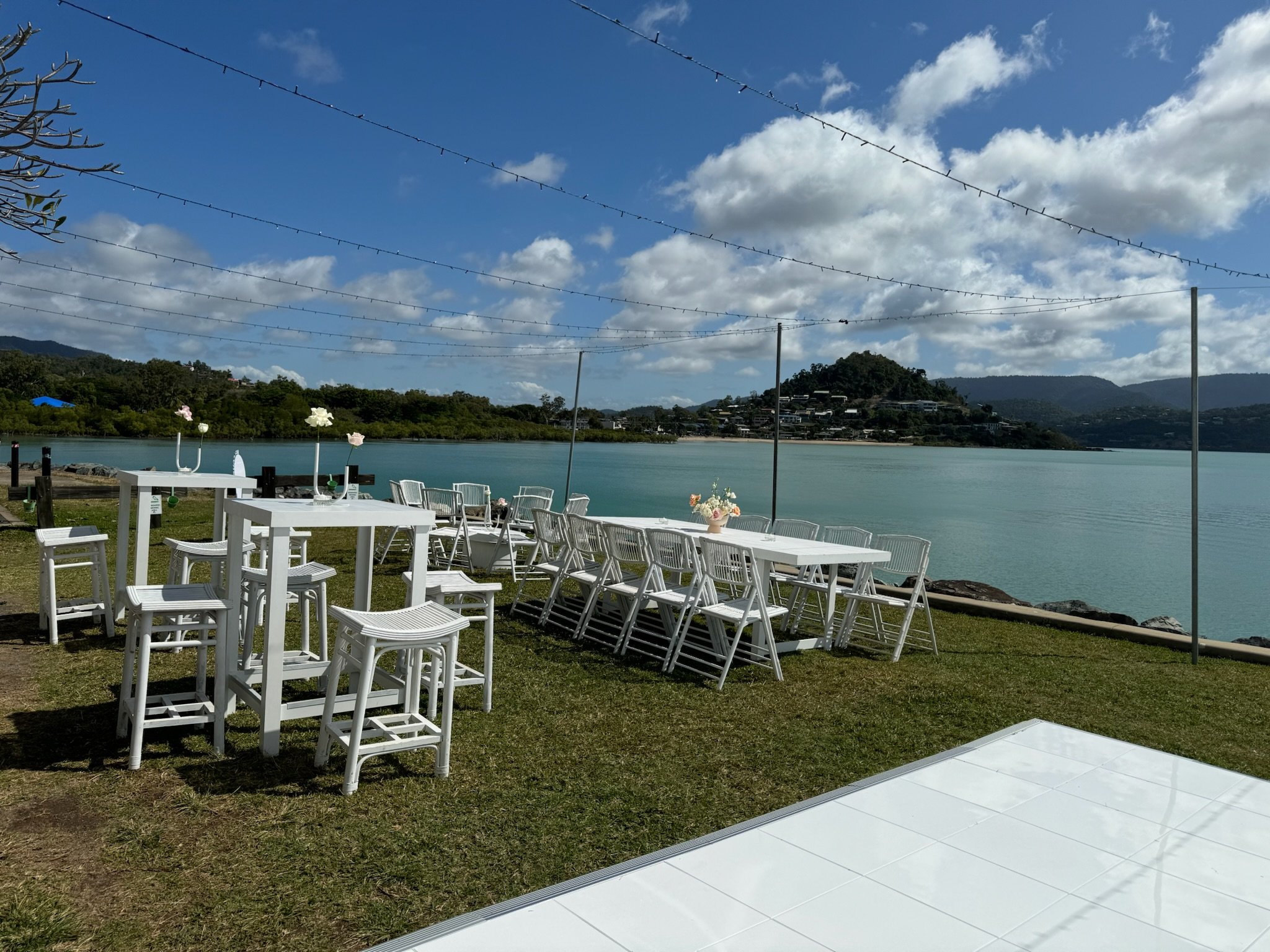 Outdoor seating area with white tables and chairs by a body of water under a partly cloudy sky, with mountains in the background.