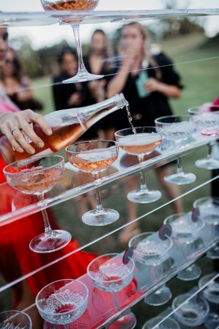 People at a party pouring champagne into coupe glasses on a multi-tiered glass serving stand.