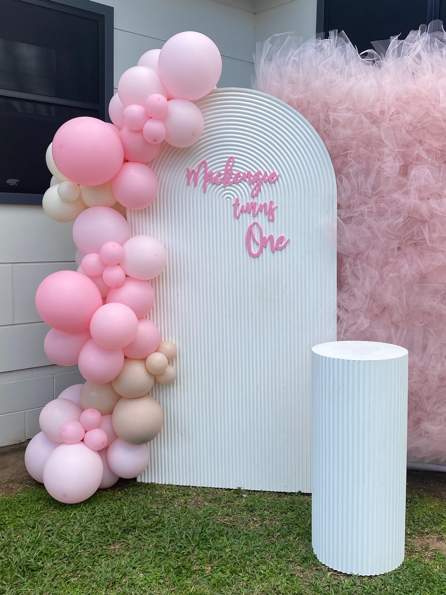Decorative setup with pink, white, and beige balloons, a white textured backdrop with pink lettering that says "Mackenzie turns One", and a pink tulle backdrop. A white cylindrical pedestal is in front of the backdrop.