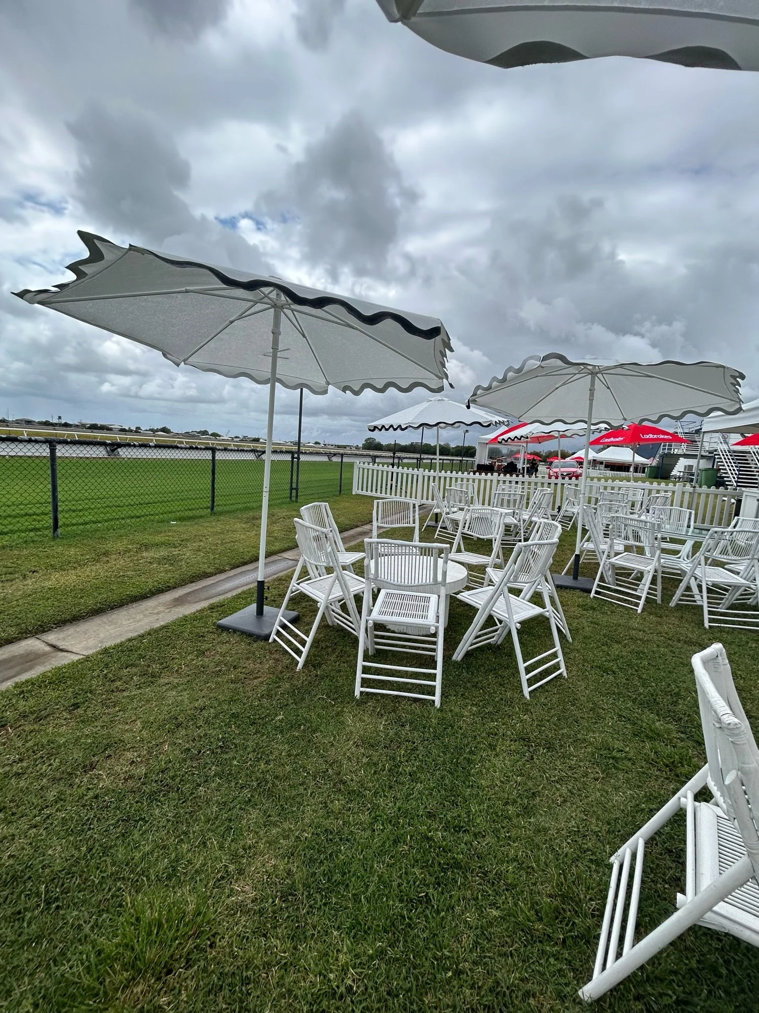 Outdoor seating area with white chairs and umbrellas on a grassy lawn, fenced with a race track in the background, overcast sky.