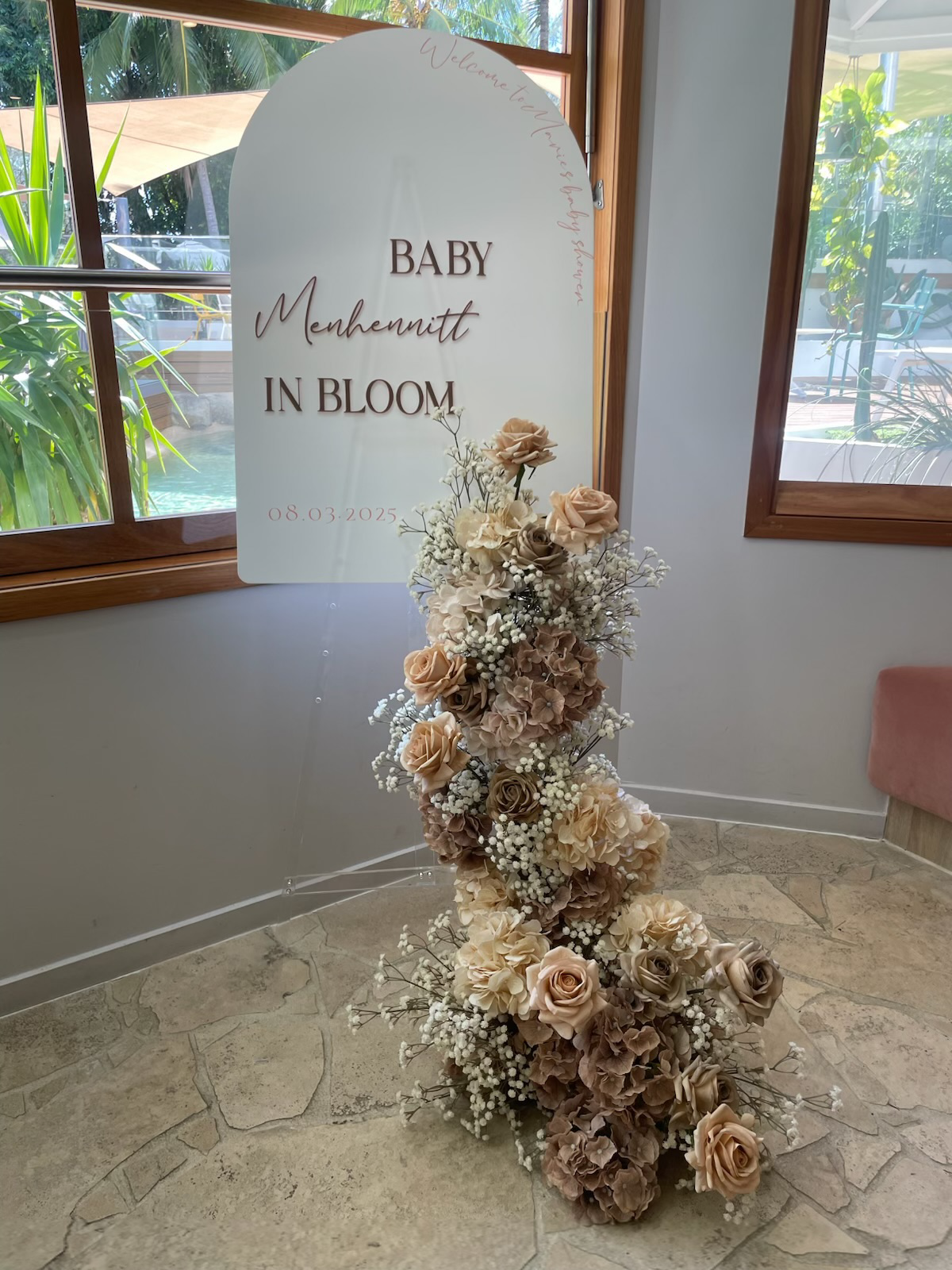 A floral arrangement of beige, white, and brown roses and baby's breath, positioned next to a white sign with brown and pink text that reads "Baby Menhennitt in Bloom" with the date 08.03.2025, inside a room with stone tile flooring and wooden window