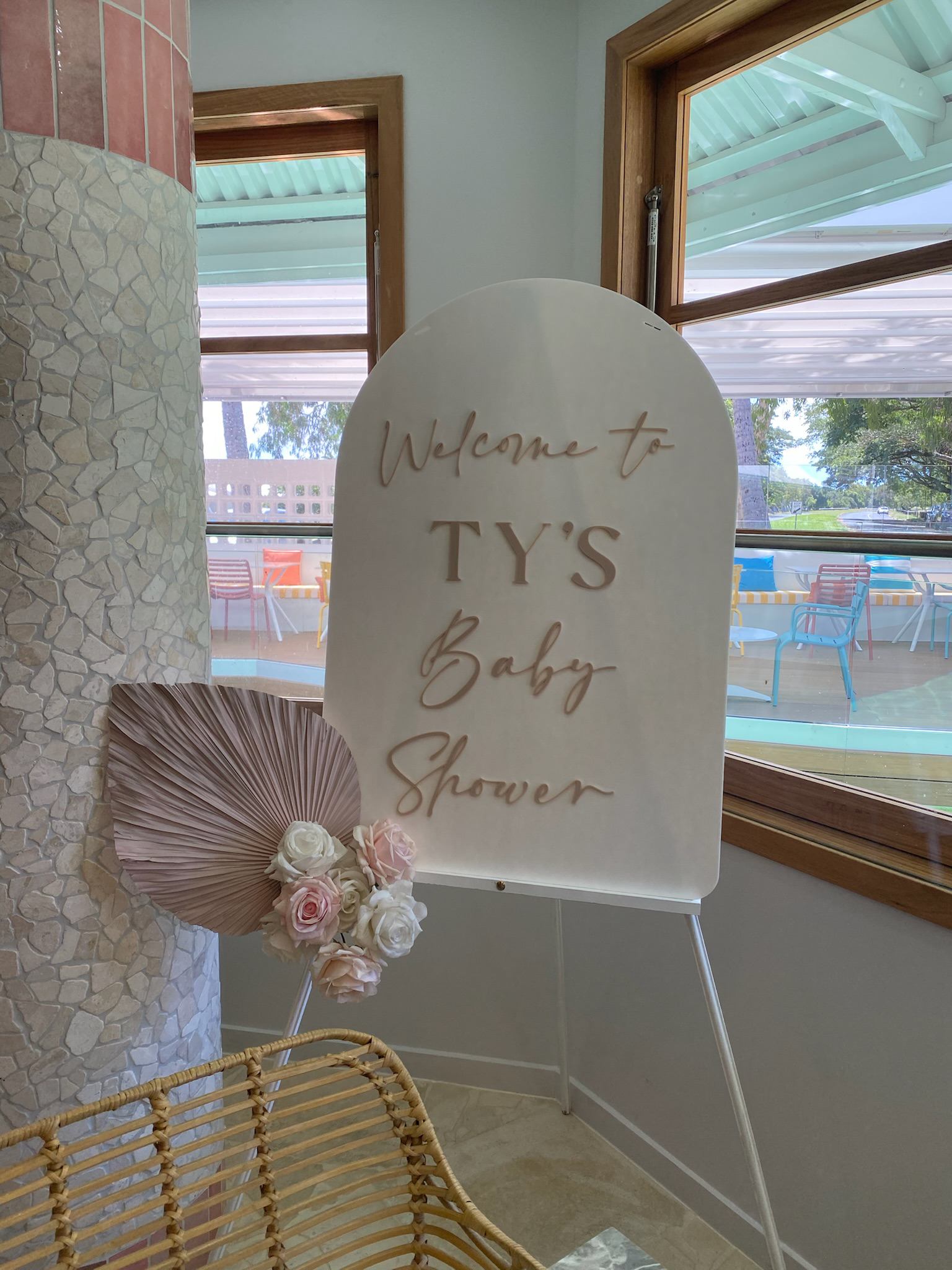 A welcome sign for a baby shower in a well-lit room with large windows showing an outdoor patio with chairs and trees.