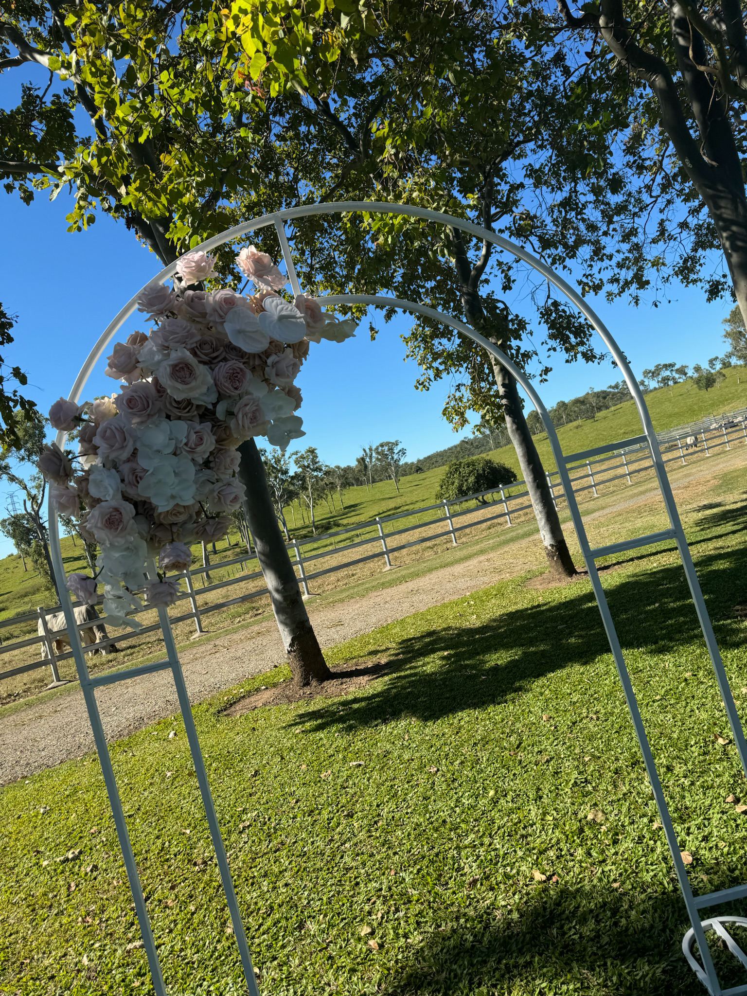 A white metal arch decorated with pink and white flowers, set outdoors on green grass with trees, a dirt path, and hills in the background.