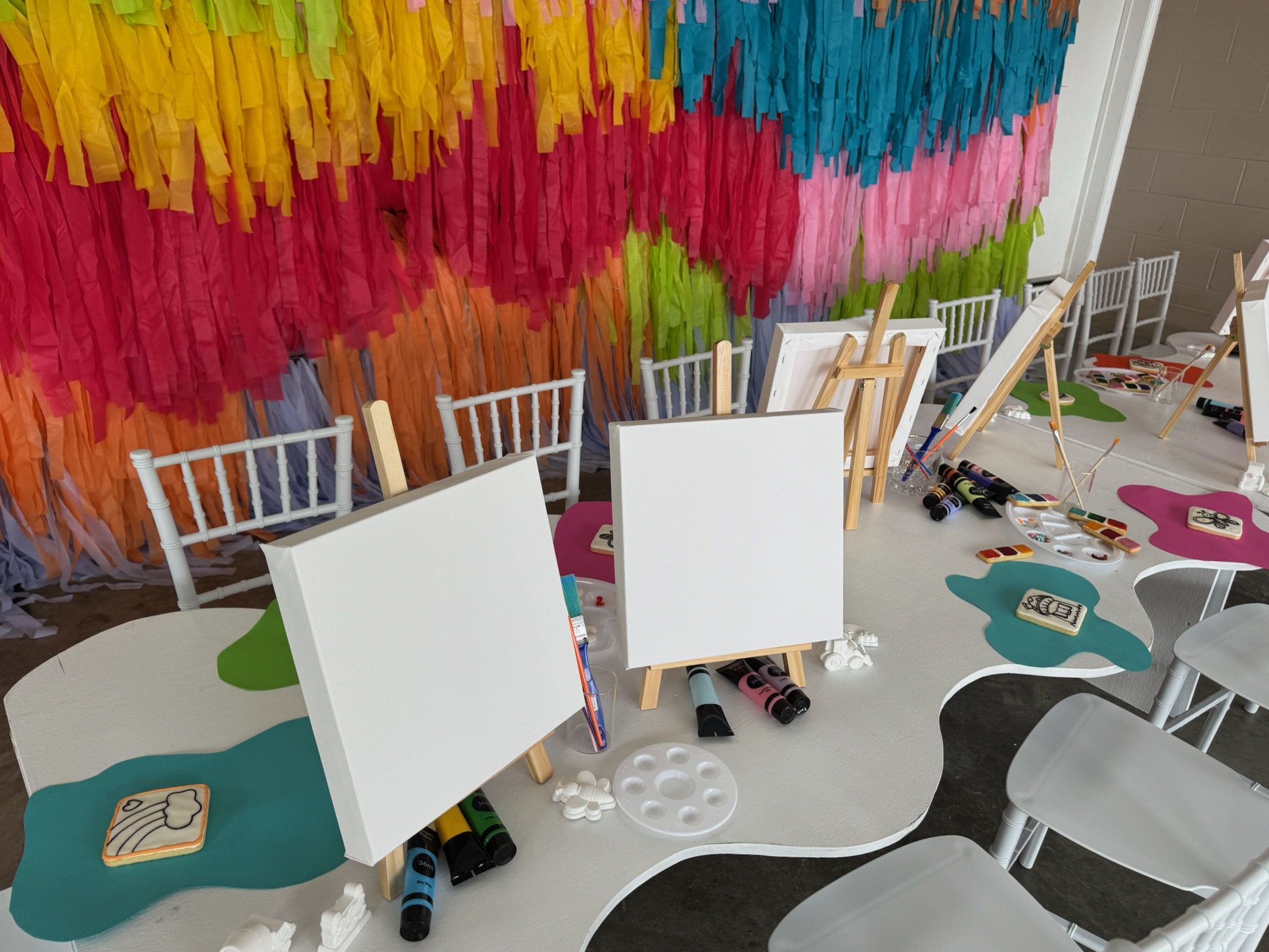 Art supplies on a white table with small canvases, paint palettes, markers, and easels, in front of a colorful tissue paper backdrop.
