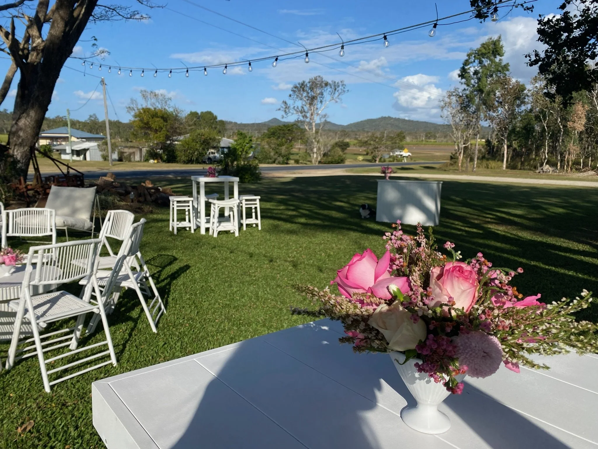 Outdoor patio with white chairs and tables, a flower arrangement in a white vase, and string lights hanging across the sky on a sunny day.