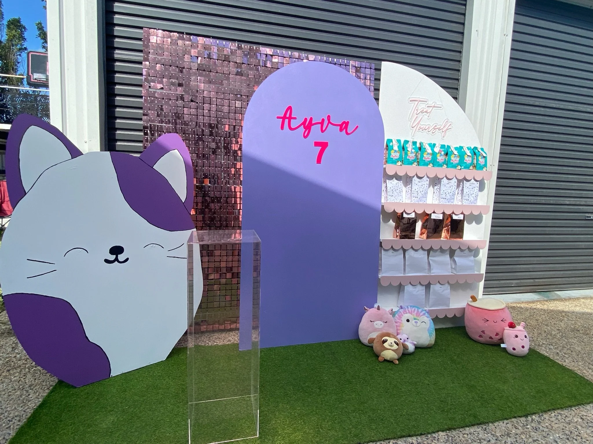 Decorative birthday display for a girl turning 7, featuring a large cartoon cat cutout, plush toys, and a purple and white backdrop with the name 'Ava' and the number 7, along with a shelf of treats and a pink pot with a stuffed animal.