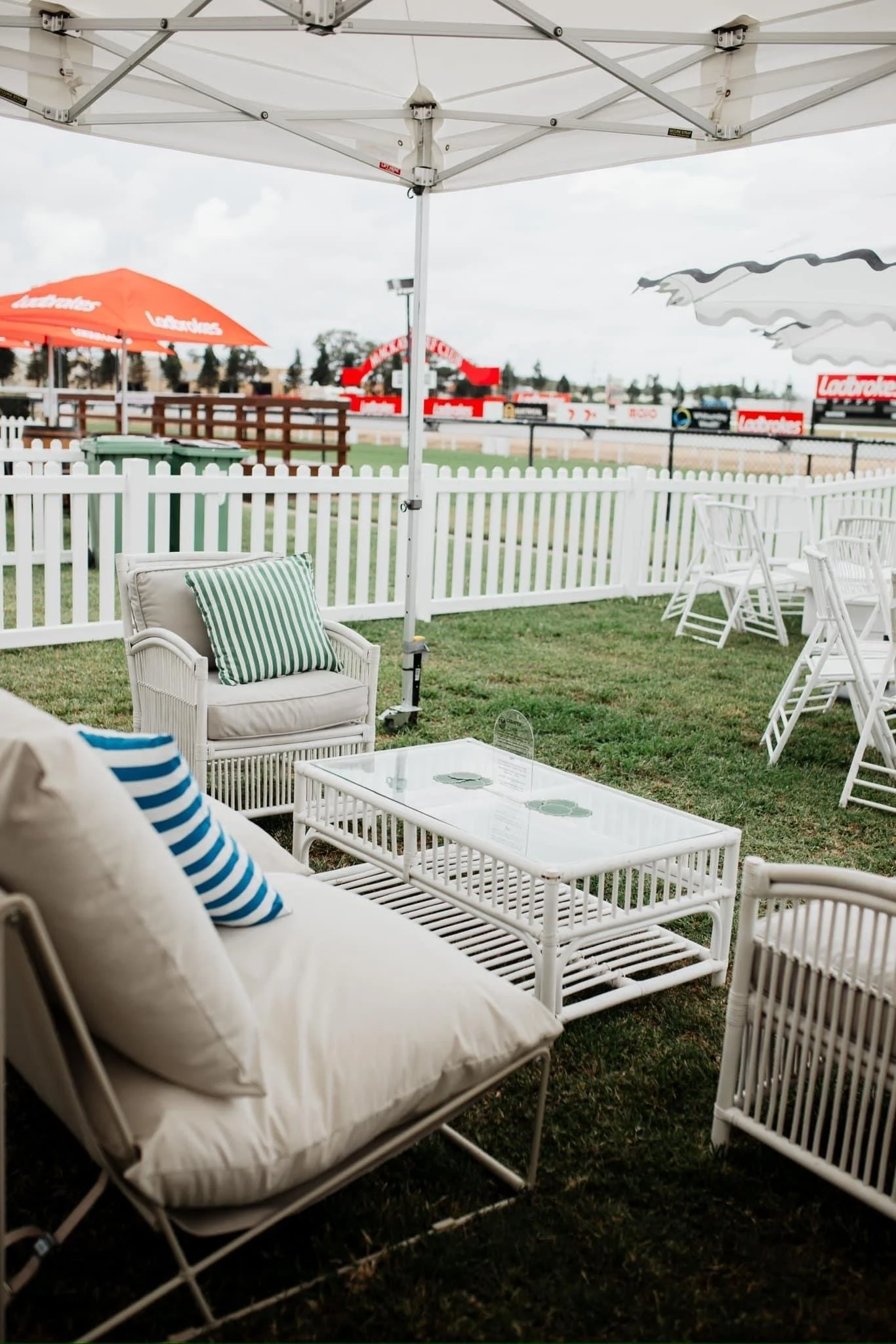 Outdoor seating area with white wicker furniture, pillows with blue and green stripes, a white coffee table with a glass top, under a white canopy. A white picket fence separates the area from a racetrack with signage and tents in the background.