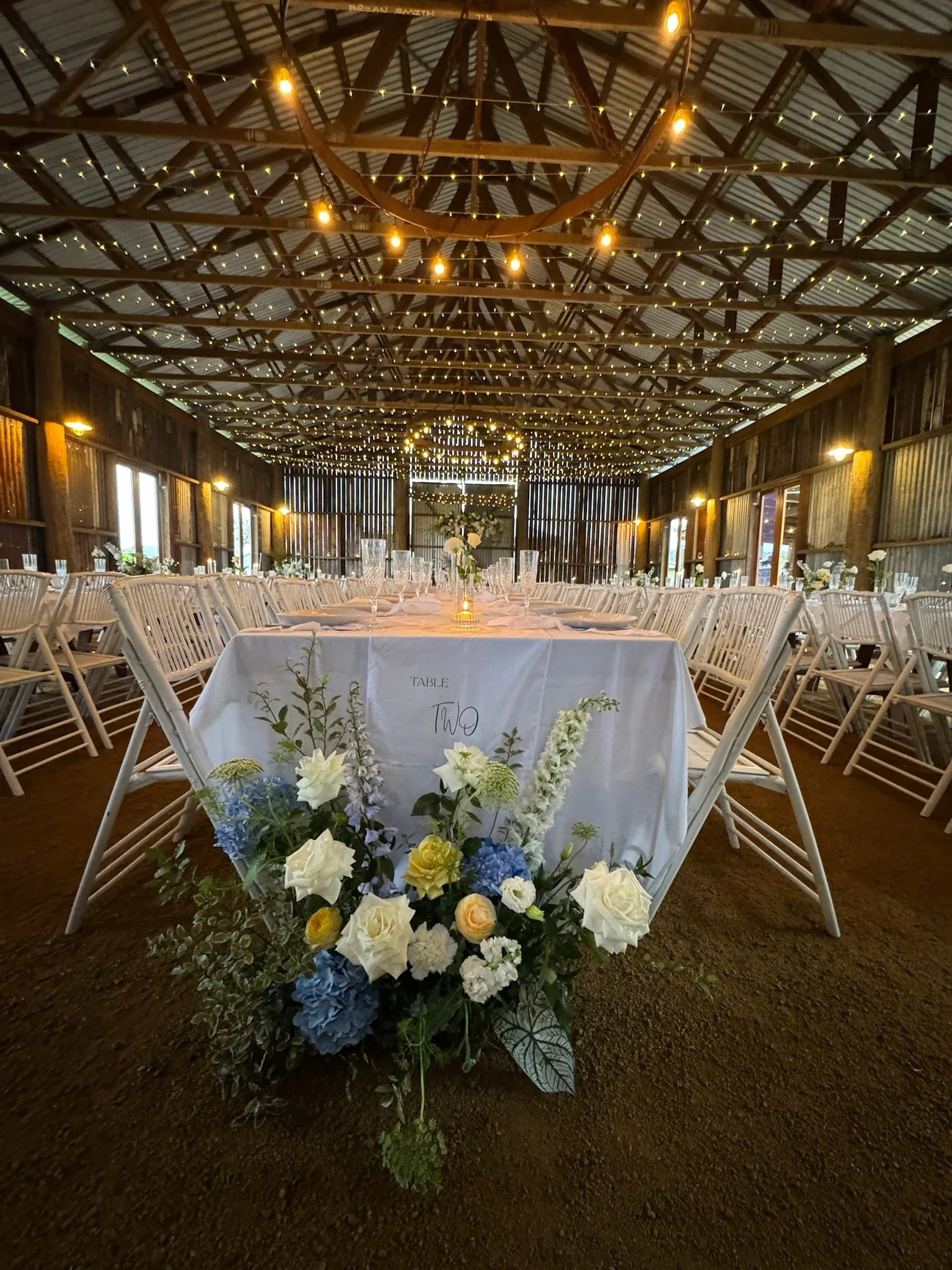 Wedding reception setup in a rustic barn with long tables, string lights, and floral centerpieces.