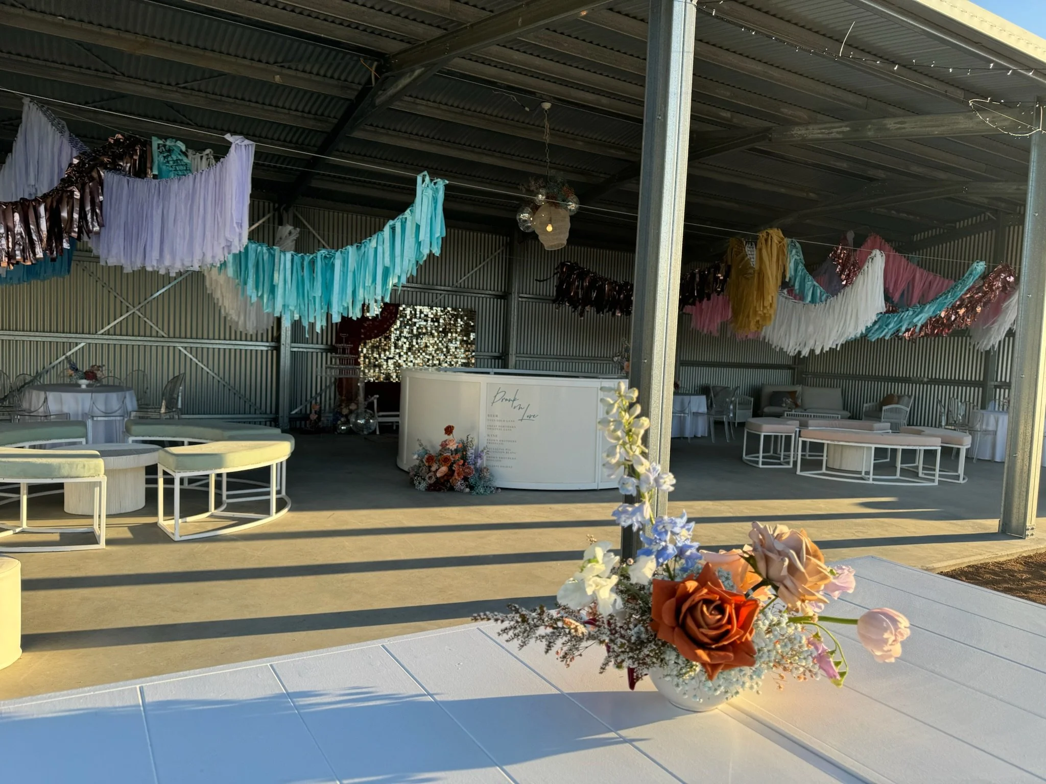 Decorated event space with colorful tassel garlands hanging from ceiling, a reception desk with floral arrangements, and seating areas including curved benches and tables.