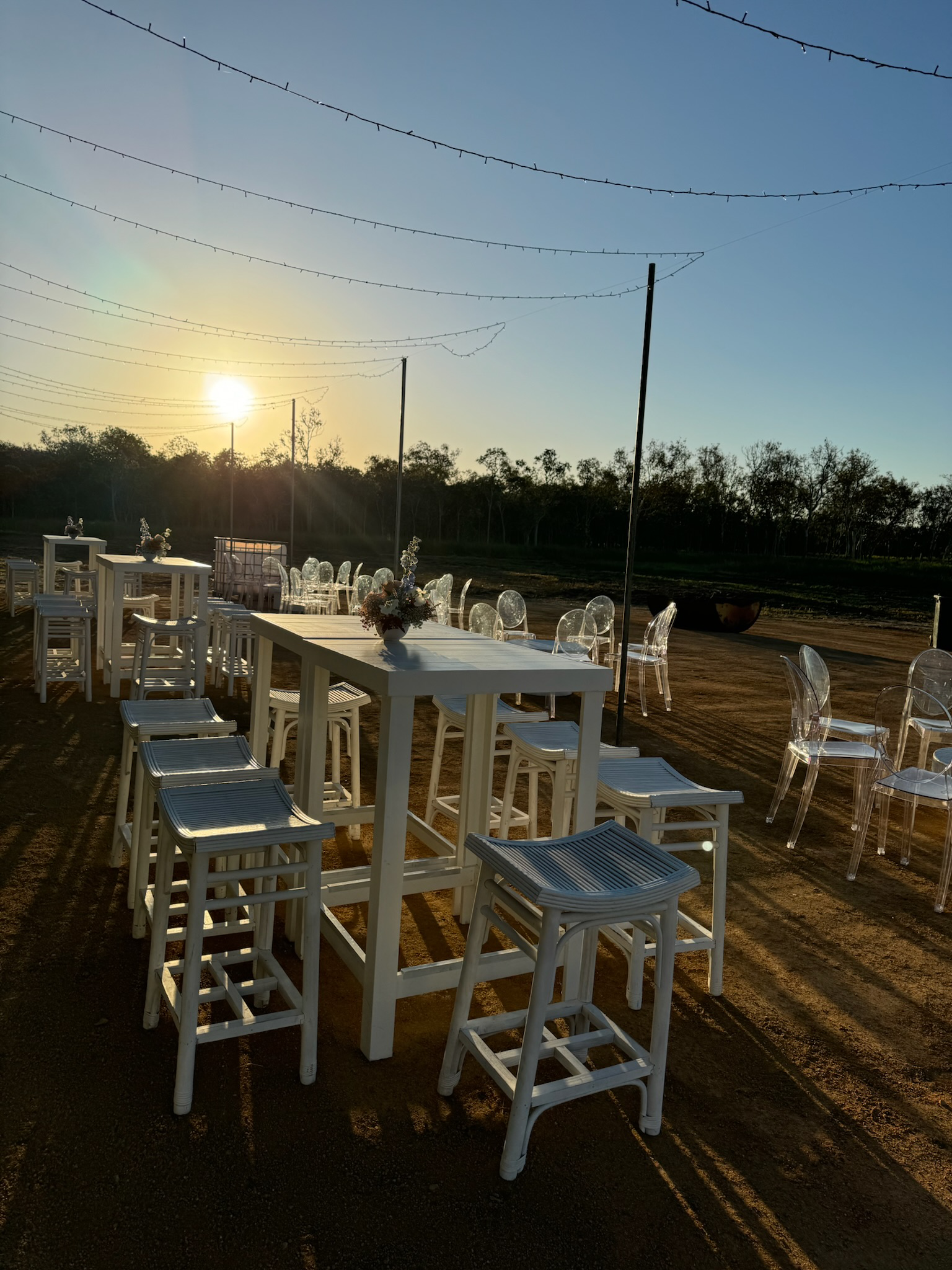 Outdoor dining setup with white tables and various chairs under string lights during sunset.