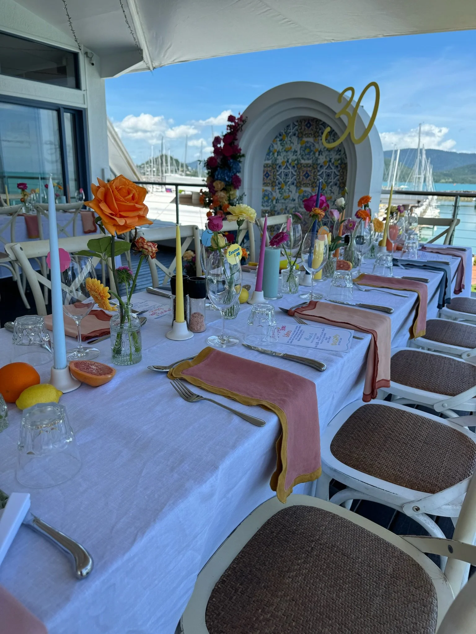 Decorated outdoor dining table set with candles, flowers, and fruit overlooking a marina with boats and hills under a blue sky. A large white arch with the number 30 in gold is in the background.