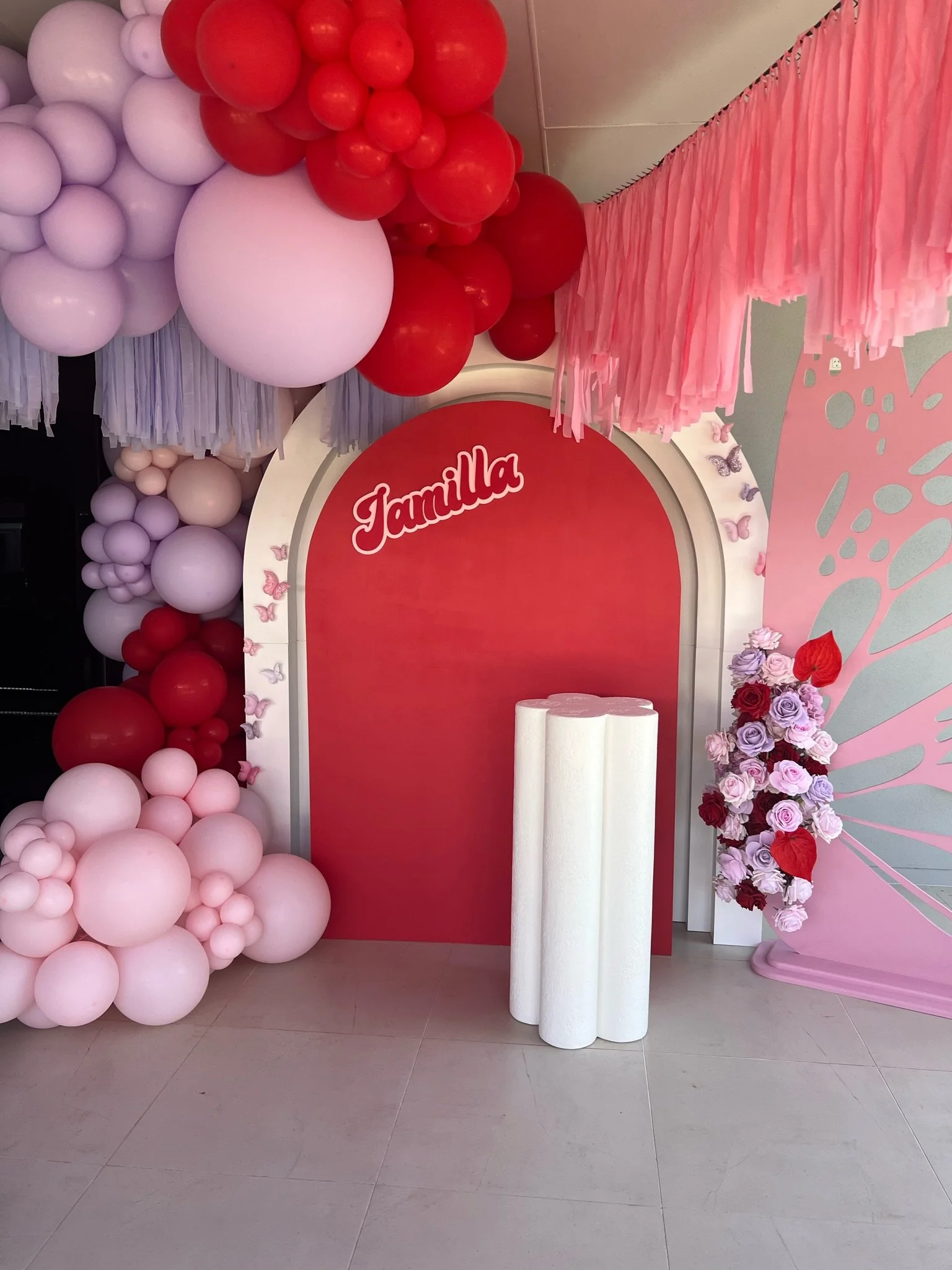 Decorative backdrop with pink and red balloons, pink paper fringe, and a floral arrangement of pink and purple roses with some red hearts, set up for a celebration with the word 'Jamilla' on a red background.