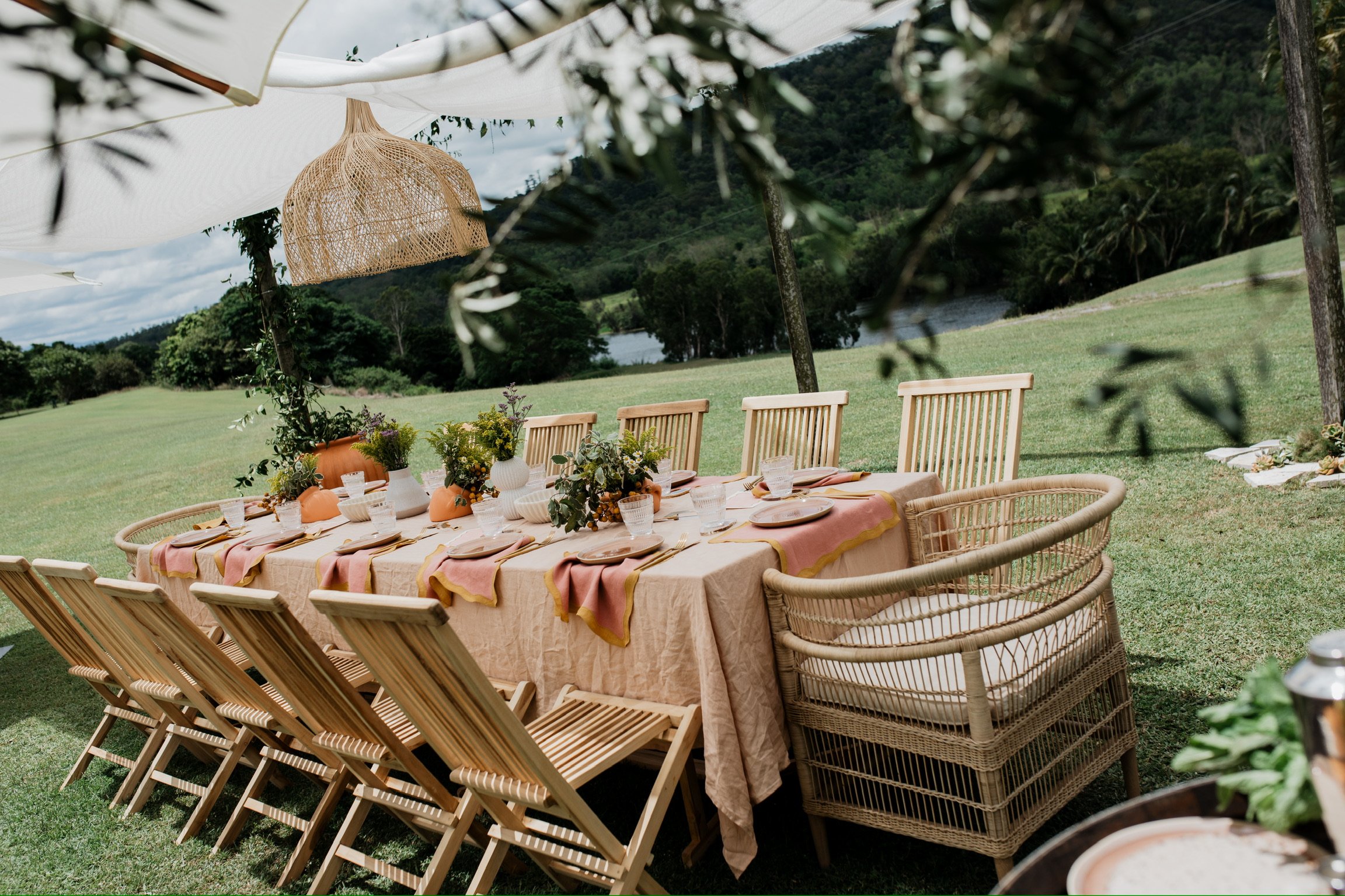 Outdoor dining setup with a long table covered in a beige tablecloth, decorated with pink and yellow table runners and floral centerpieces in vases, surrounded by wooden and wicker chairs on a grassy field with trees and mountains in the background.
