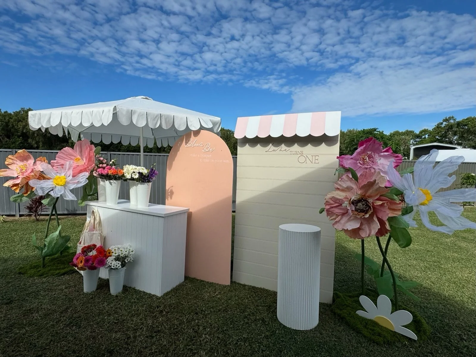 Outdoor scene with decorative floral setup, including large paper flowers, small potted flowers, and a white canopy, against a backdrop of a blue sky with scattered clouds and a fence.