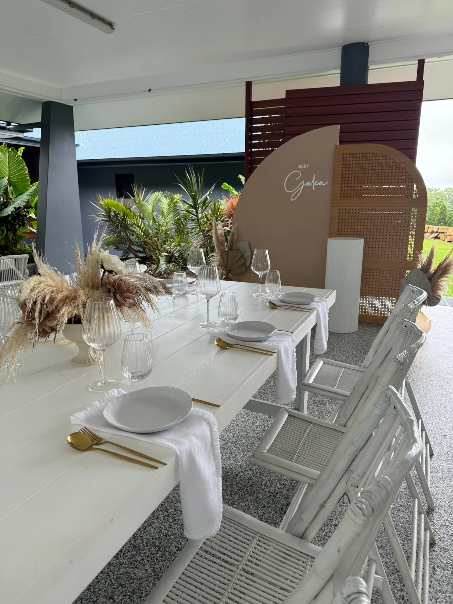 Elegant outdoor dining table set with white plates, gold cutlery, wine glasses, and floral centerpieces, in front of a decorative backdrop with the words "Baby Galea".