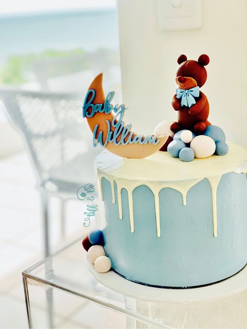 A two-tiered cake with white and blue icing, decorated with colorful balls and a teddy bear topper. A moon-shaped cake topper with the words "Baby William" written on it is attached to the cake.