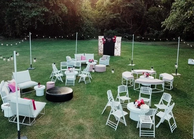 An outdoor event setup with white chairs and tables, pink cushions, string lights, and a floral backdrop with a chalkboard, on a grassy lawn surrounded by trees.